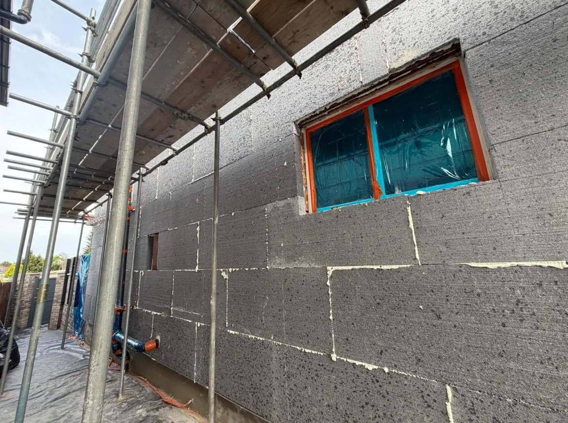 Construction site showing a building with concrete block walls, scaffolding, and a window covered with blue plastic.