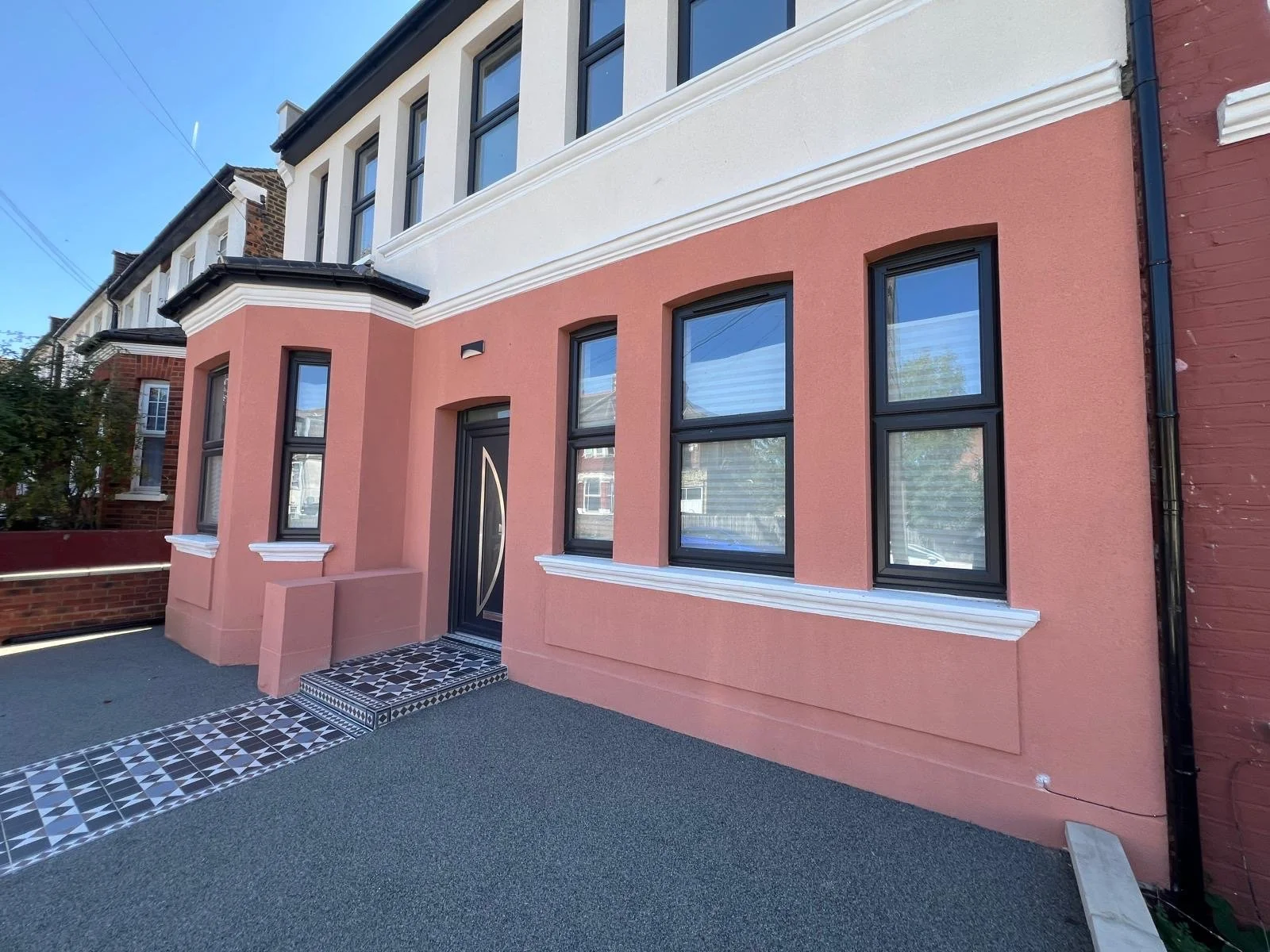 Pink and white multi-story residential building with black framed windows and a black front door, featuring a tiled entryway and a textured ground surface.
