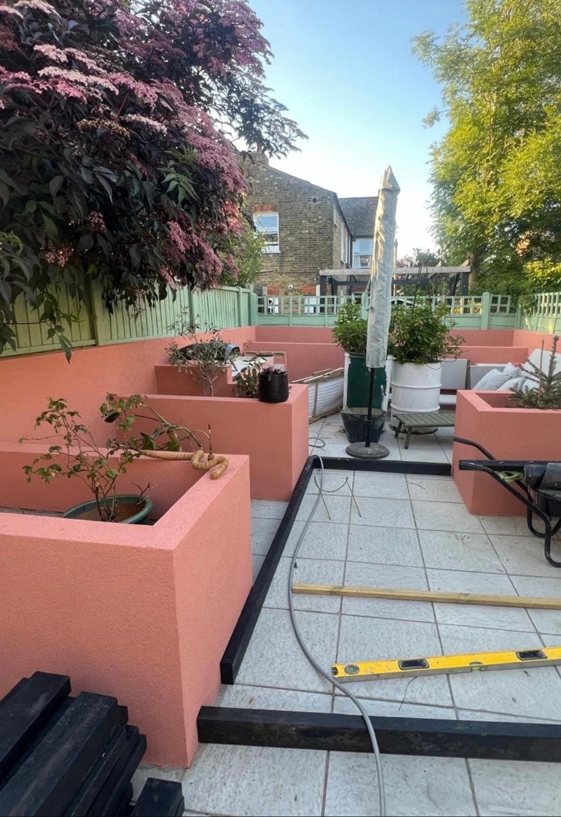 A rooftop terrace with pink and green planters, potted plants, outdoor furniture, a closed umbrella, and construction tools and materials, including a level and hoses, against a backdrop of trees and neighboring buildings.