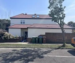 A residential house with a sloped roof, solar panels, and a garage, with three trash bins outside, a tree, and a street in front.