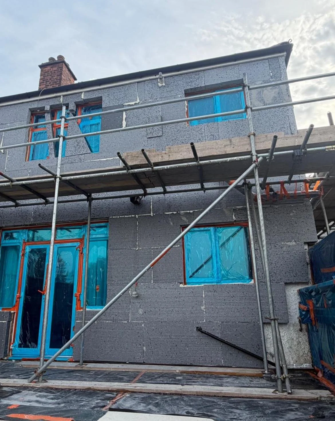 Construction site with a two-story building covered in gray insulation material, surrounded by scaffolding, with blue windows and doors wrapped in protective plastic.