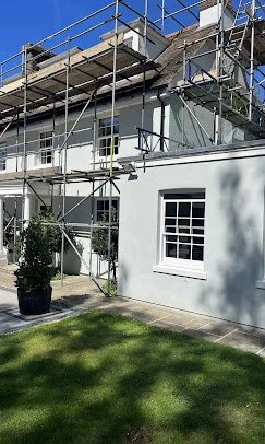 A two-story white house with scaffolding around it, indicating construction or renovation work, with a lawn and potted plant in the foreground.