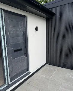 Exterior corner with a black door and black wall paneling, white wall, and concrete paving.