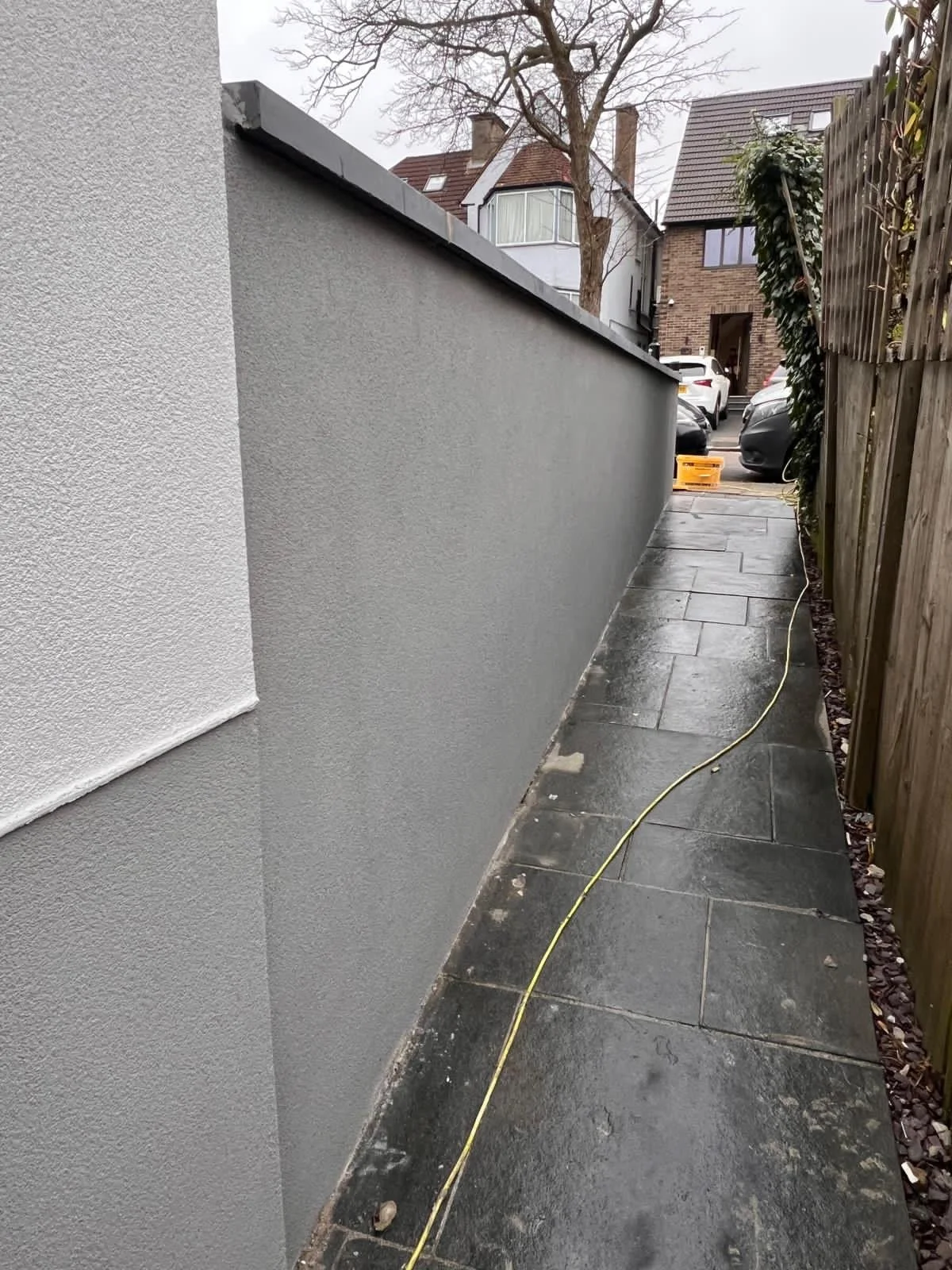 A narrow sidewalk next to a gray building wall, with a wooden fence on the right and a yellow electrical cord running along the ground, wet pavement, and parked cars in the distance.