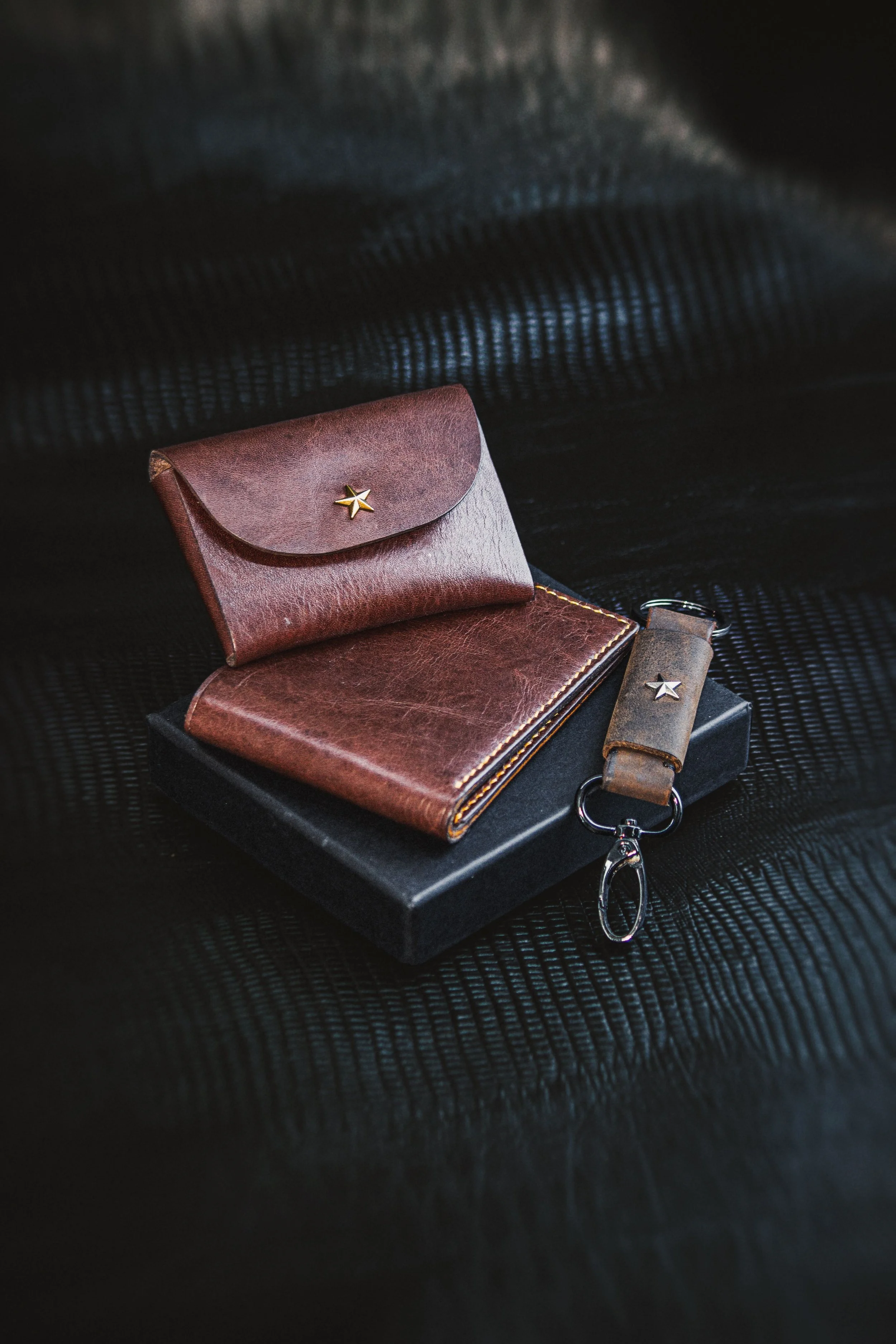 A brown leather wallet with a gold star emblem, a matching keychain, and a gray wallet, placed on a black textured surface.