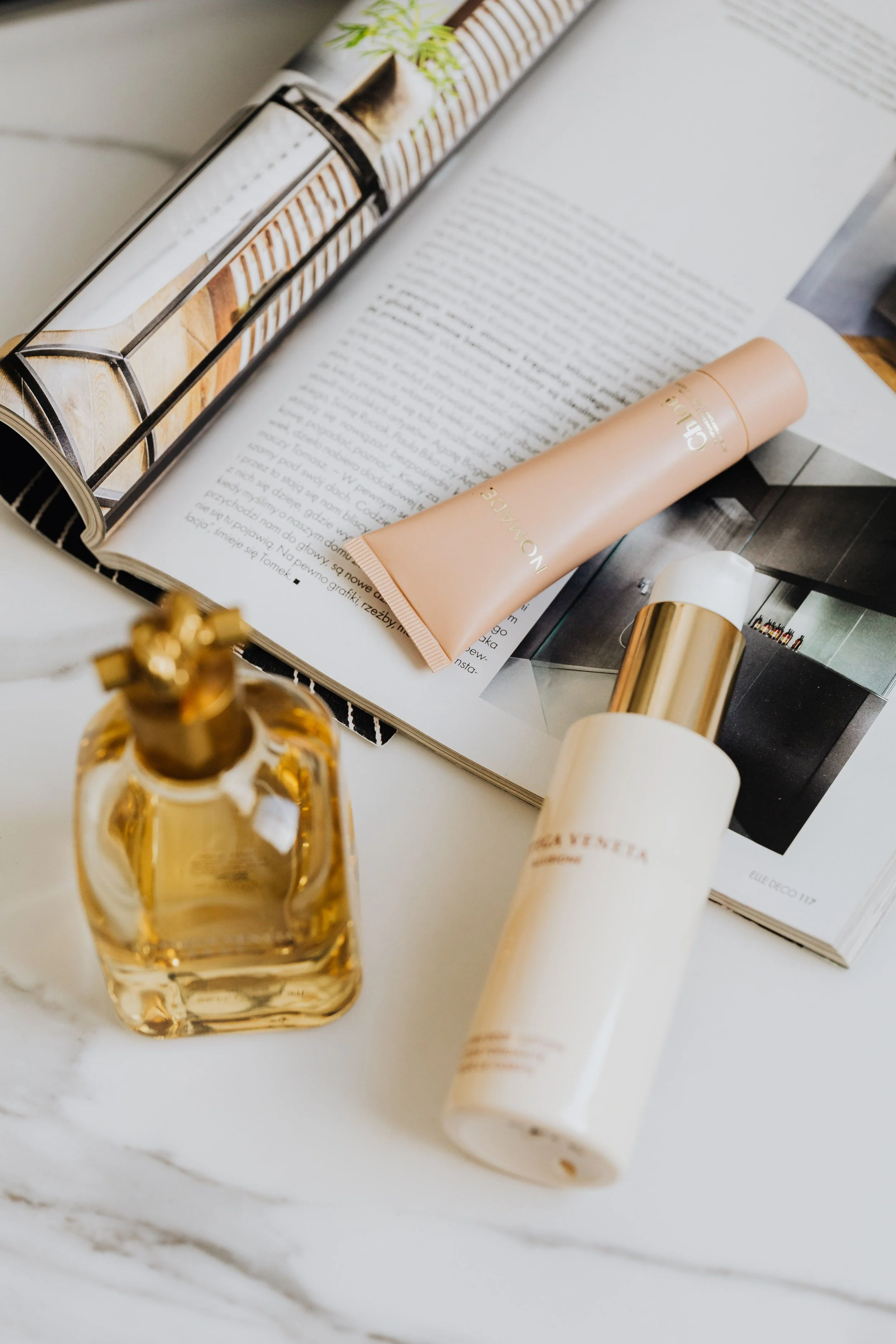 A flat lay of beauty products on a white marble surface, including a gold perfume bottle, two tubes of cream, and a magazine open to an interior design page.