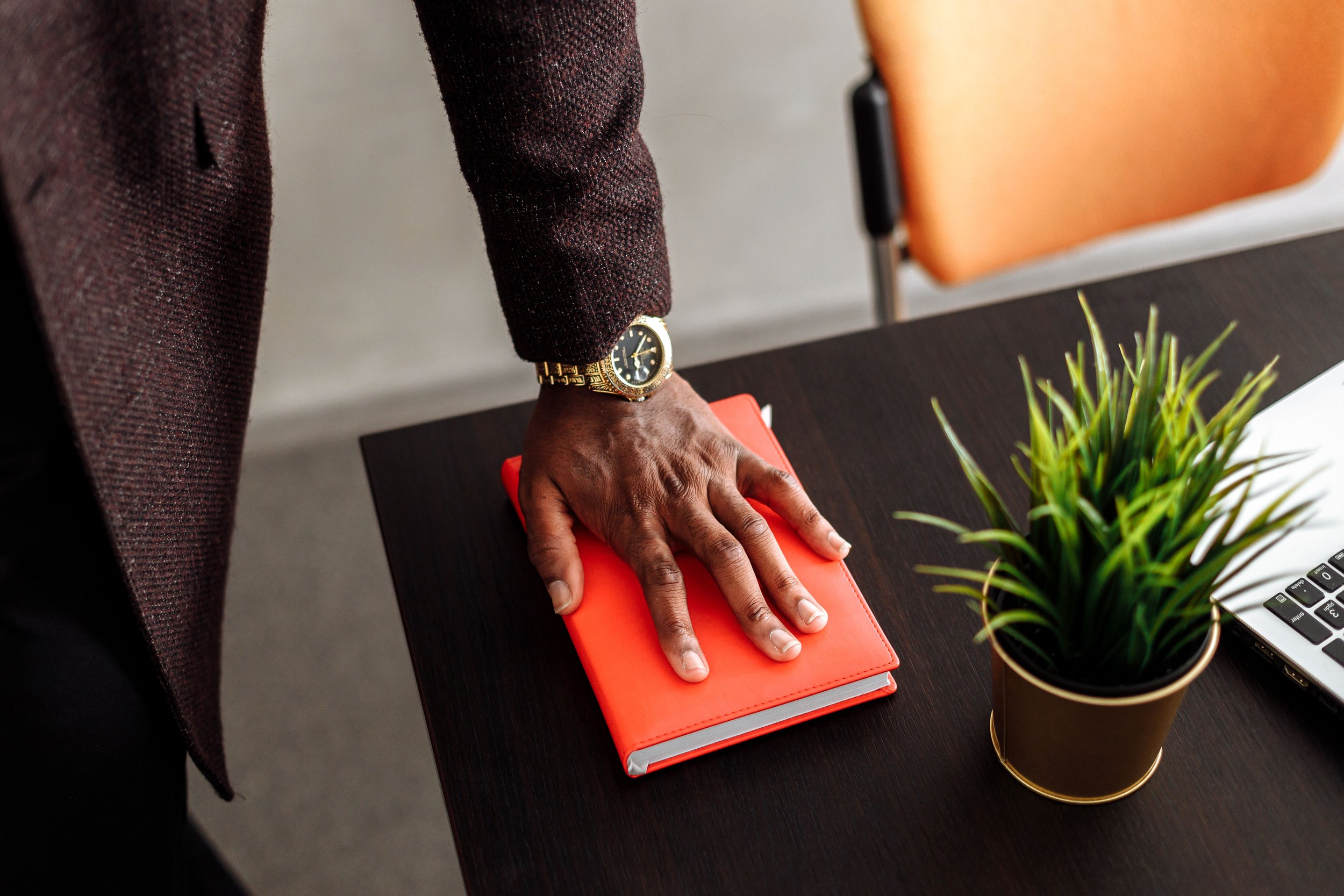 Person wearing a watch, placing a hand on a red notebook or planner on a dark wood desk. There is a green potted plant and a black and white keyboard nearby. The person is dressed in a dark, textured blazer.