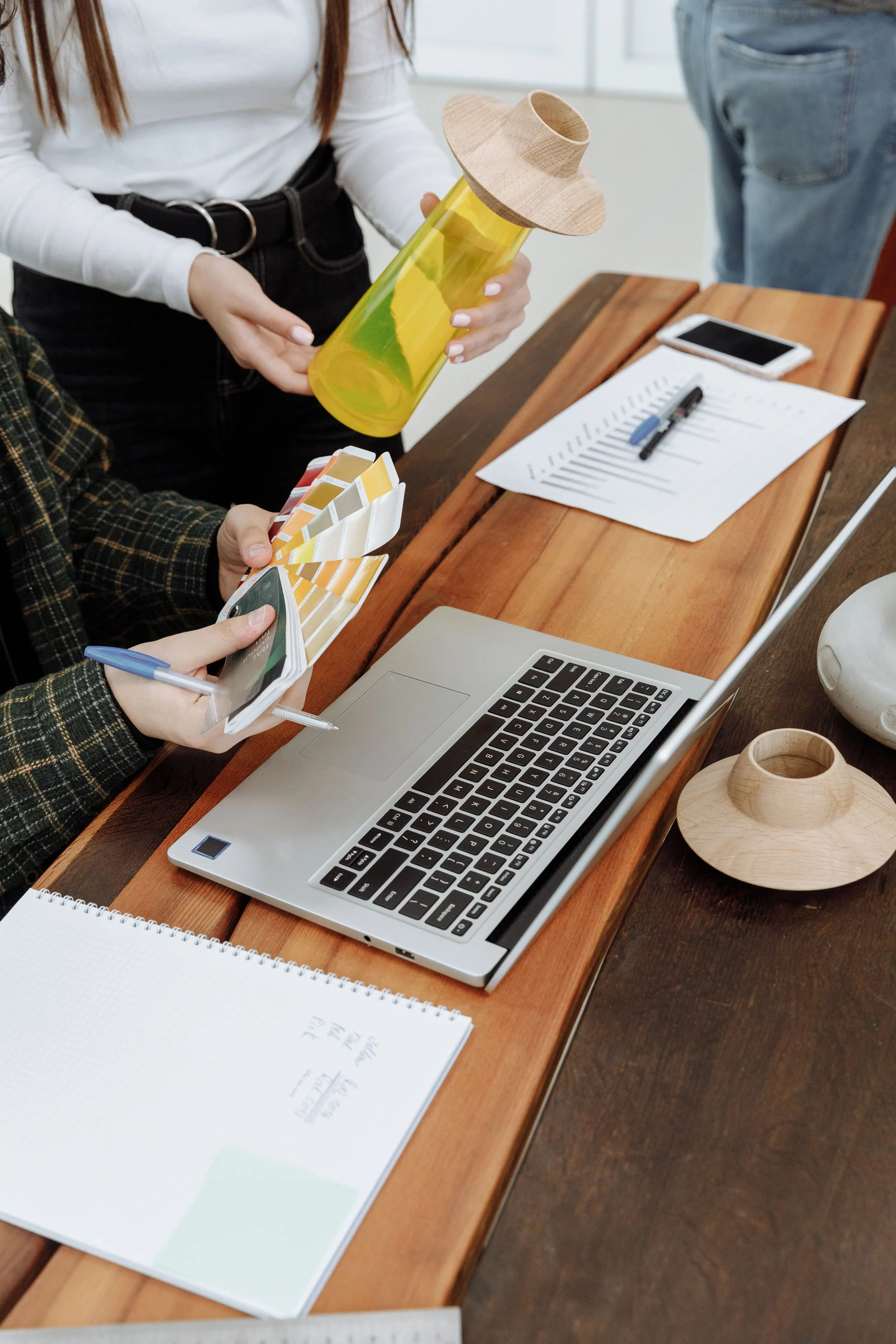 A person holding a yellow water bottle with a wooden hat-shaped lid in a workspace with a laptop, notebook, pens, and a smartphone on a wooden desk.