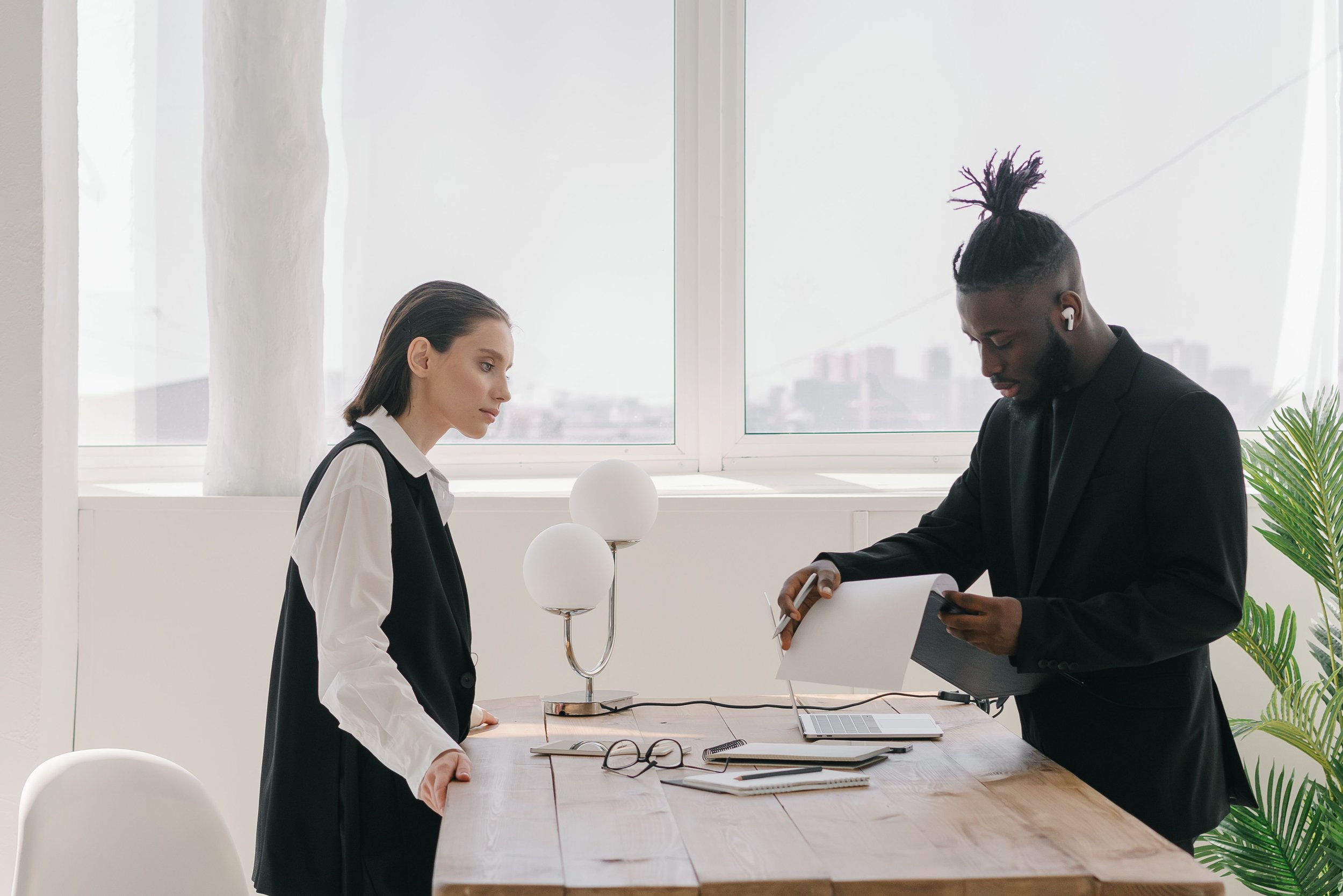 A woman and a man dressed in business attire are standing at a table in a bright office space, engaged in conversation, with the woman looking at the man and the man looking down at a folder. There is a lamp, a laptop, a pair of glasses, and some notebooks on the table, and a large window with urban buildings outside in the background.