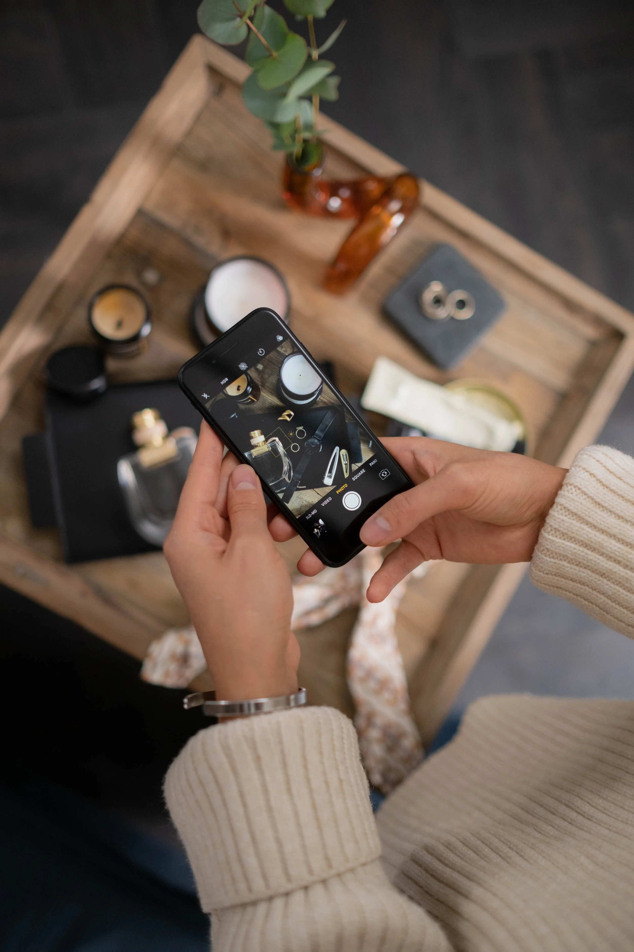 Person taking a photo of a wooden tray with candles, a small vase with greenery, jewelry, and skincare products, wearing a beige sweater and a bracelet.