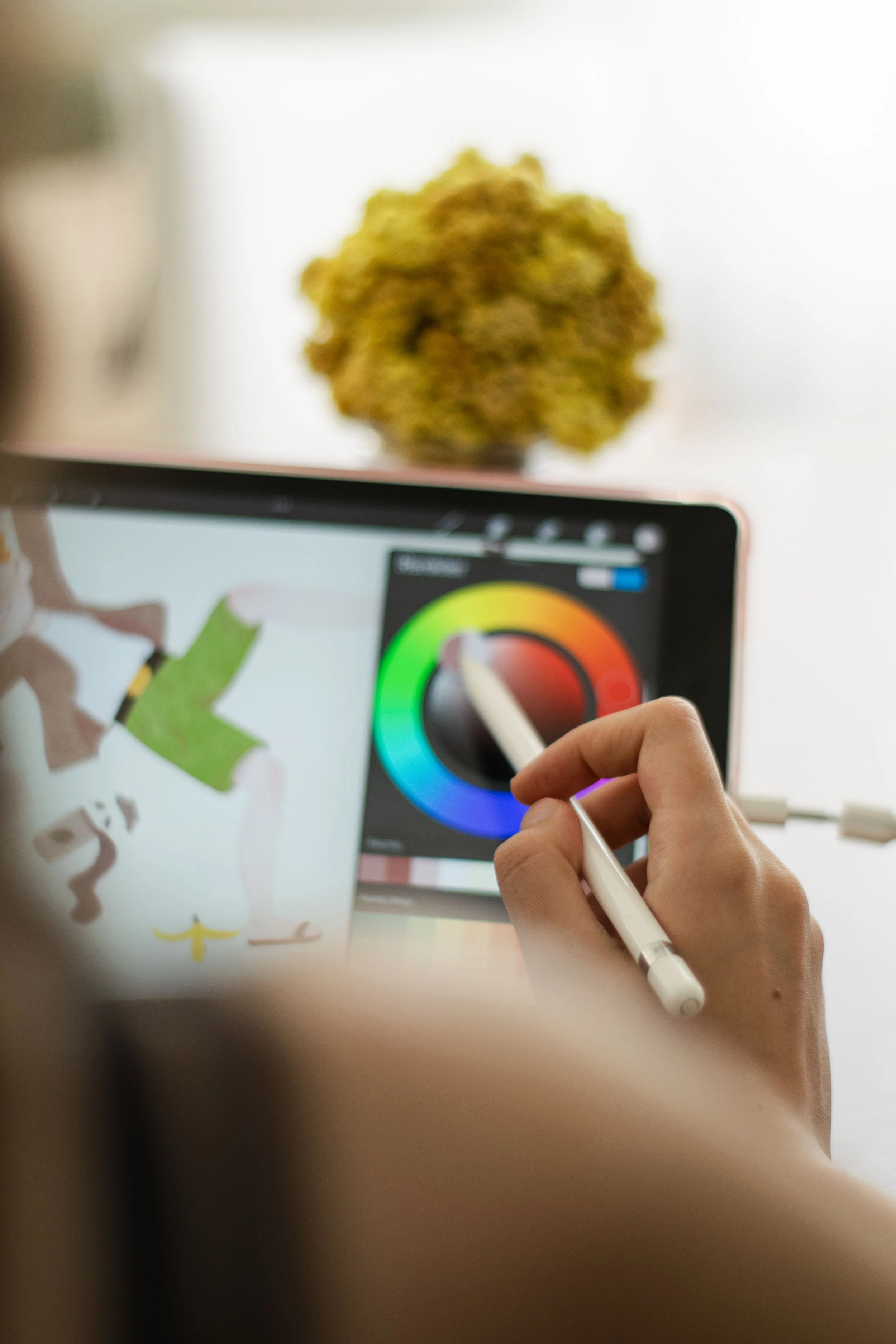 Close-up of a hand using a stylus on a tablet screen displaying a color wheel, with blurred artistic sketches and a green character in the background.