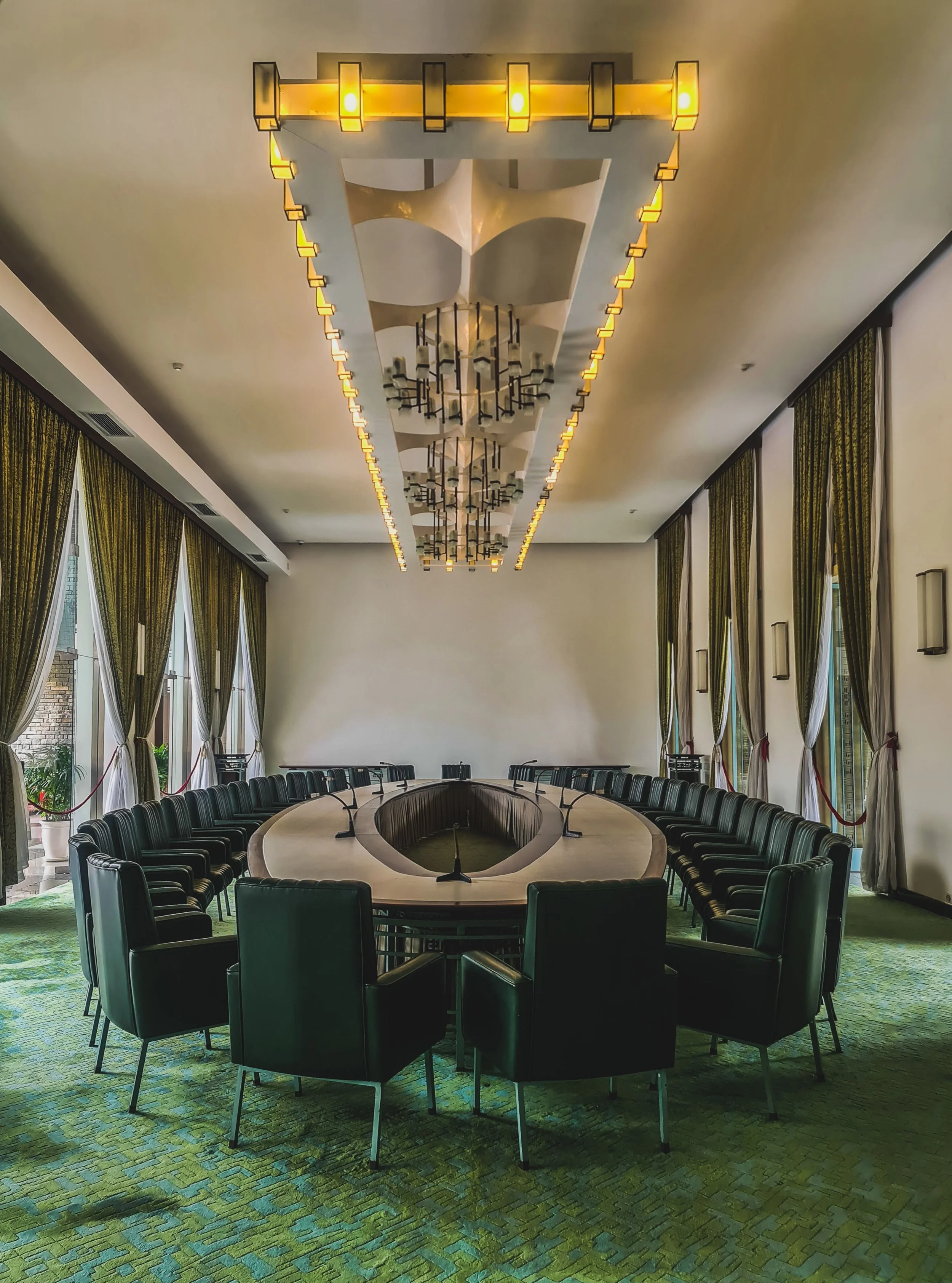 A large, U-shaped conference table surrounded by black leather chairs in a meeting room with green patterned carpet, tall windows with green and yellow curtains, and a modern ceiling chandelier with multiple light fixtures.