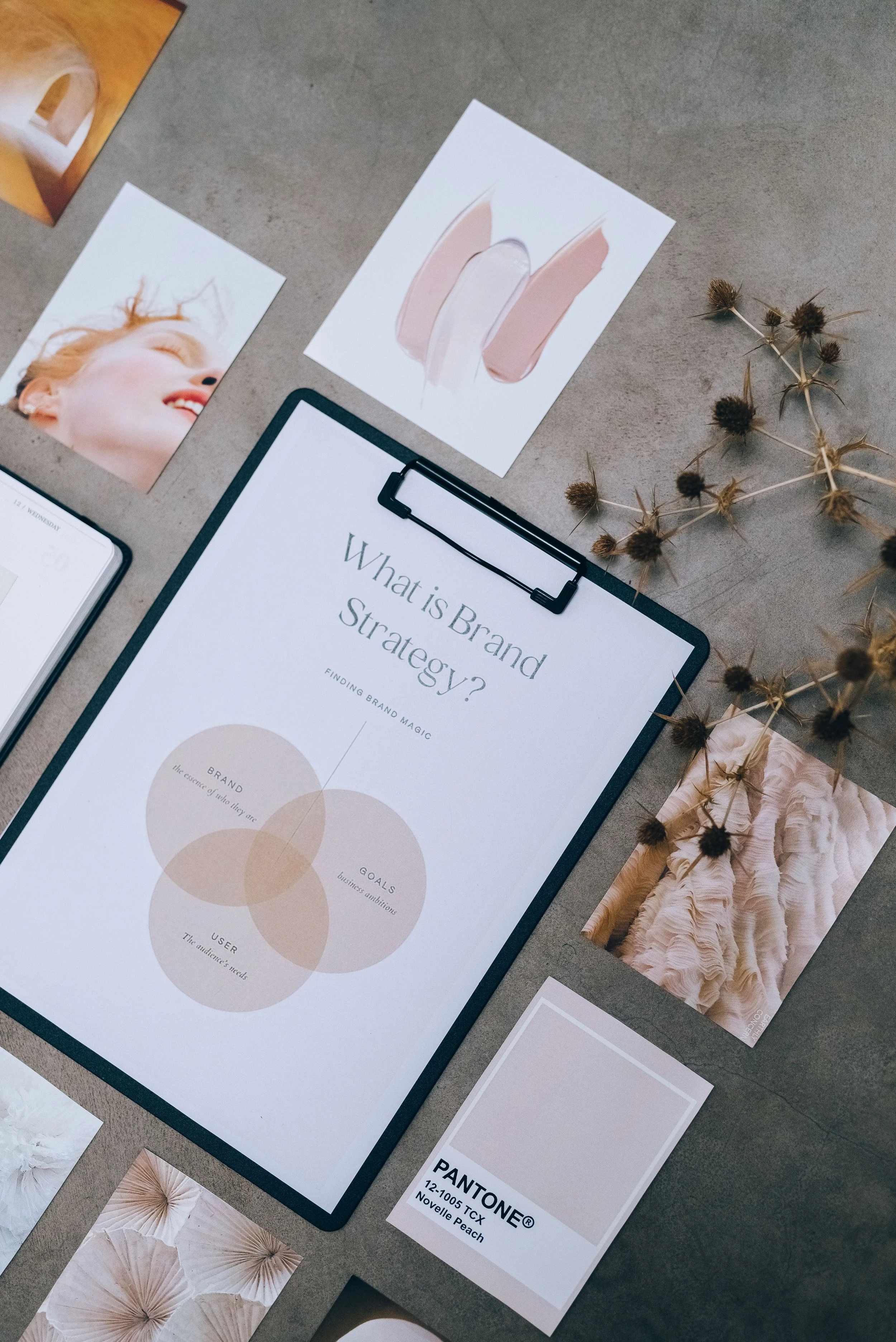 A flat lay of various design and branding materials including a printed image of dried flowers, a woman smiling, a color palette, and a presentation slide titled 'What is Brand Strategy?'. There is also a Pantone color swatch labeled 'Novelle Peach' and some dried thistle stems.