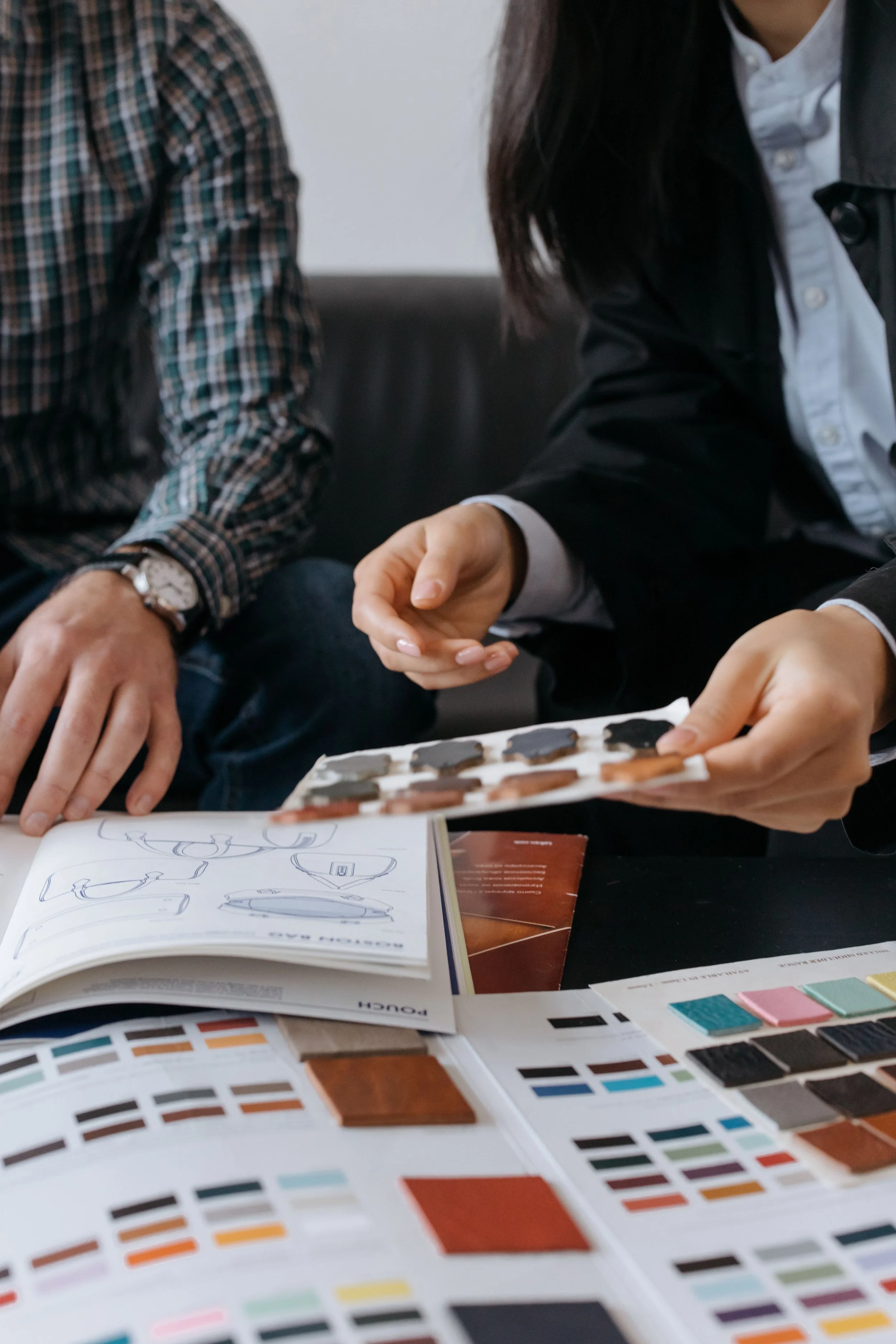 Two people are sitting at a desk, looking through design or color samples, with paint swatches, color palettes, and an open design book on the table.