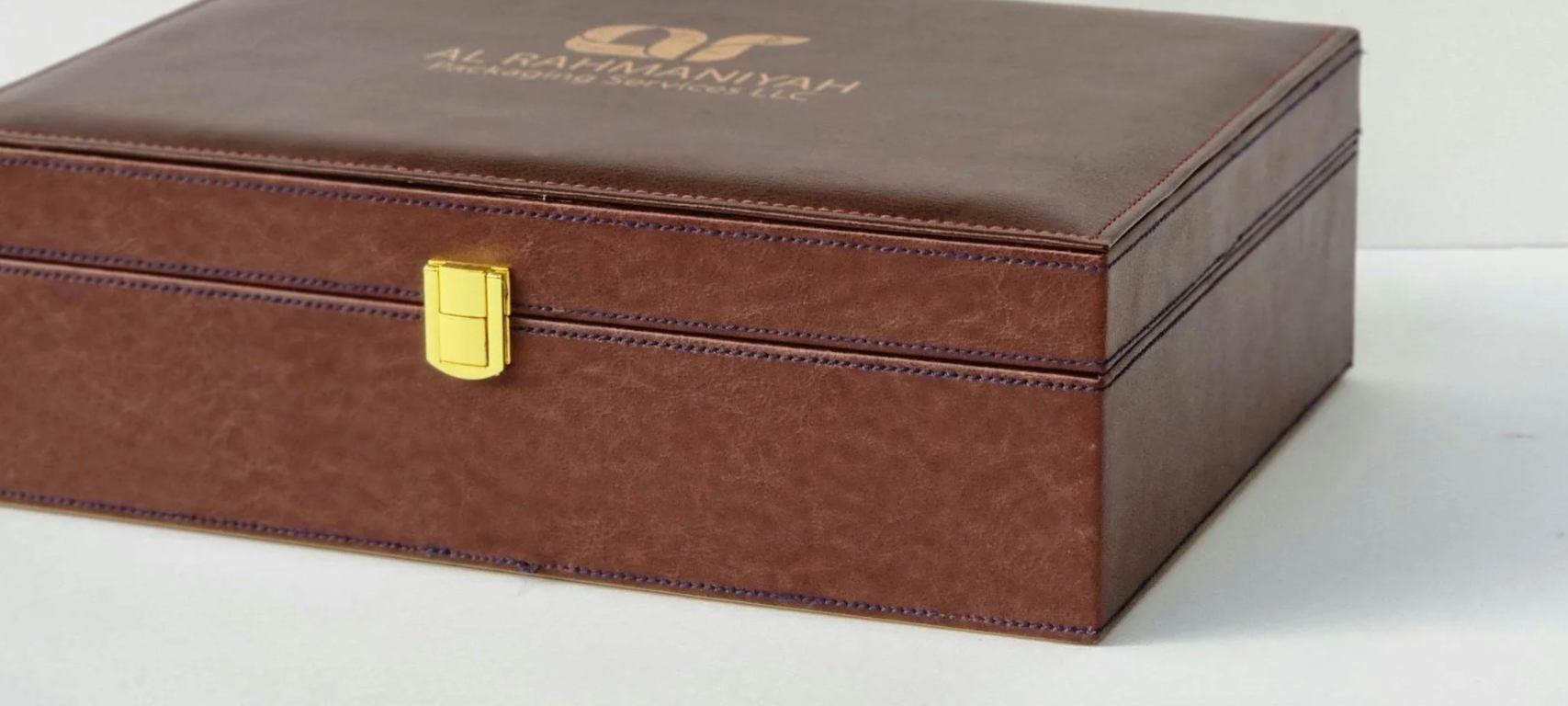 A rectangular brown leather box with gold latch and blue stitching, displaying the logo 'AL-RAHMANIYAH AL-ABYADH & CO. LLC' on the top.