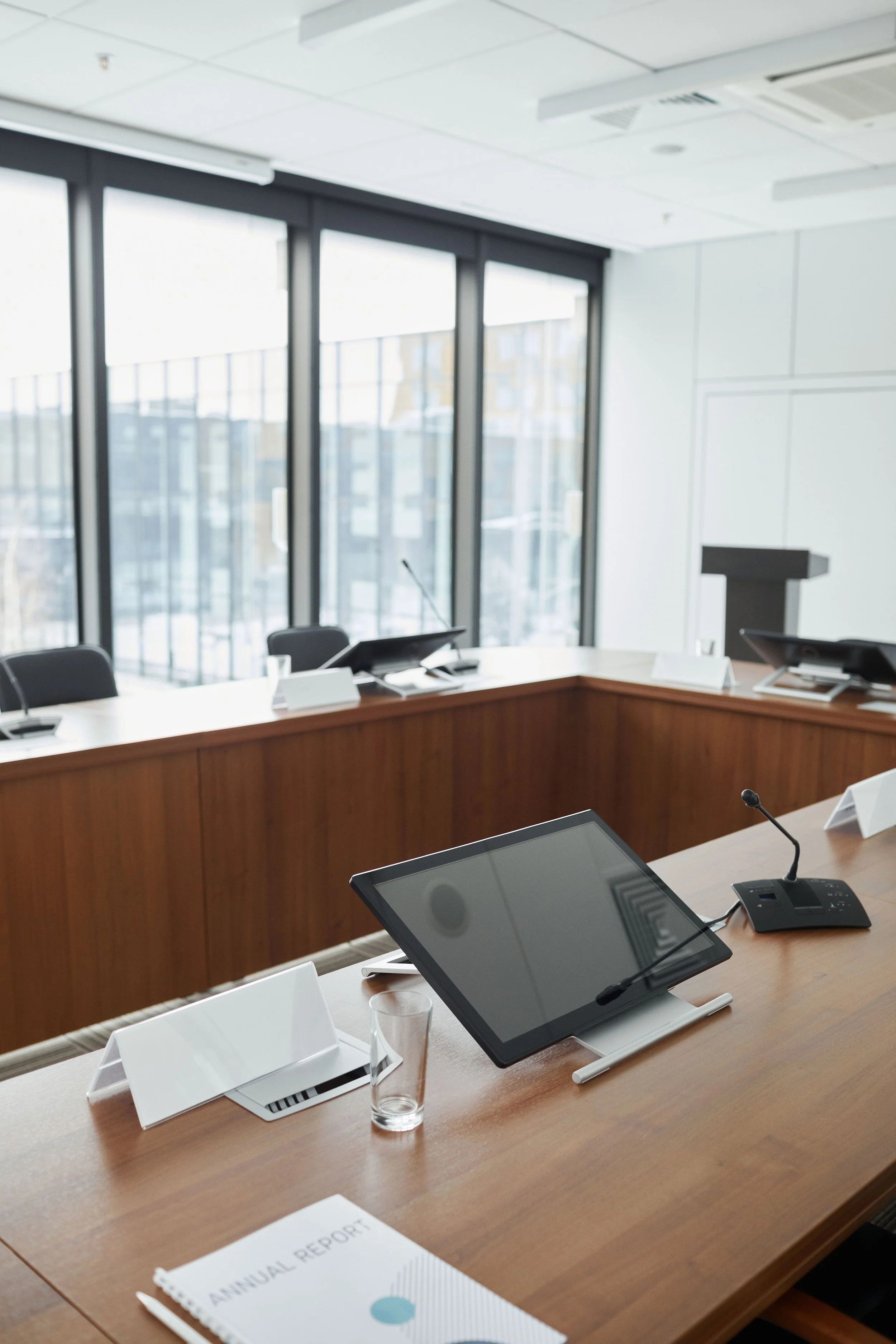 A conference room with a long wooden table, several black teleconferencing devices, and digital tablets on stands. There is a glass of water, papers, and pens on the table. Large windows in the background let in natural light.