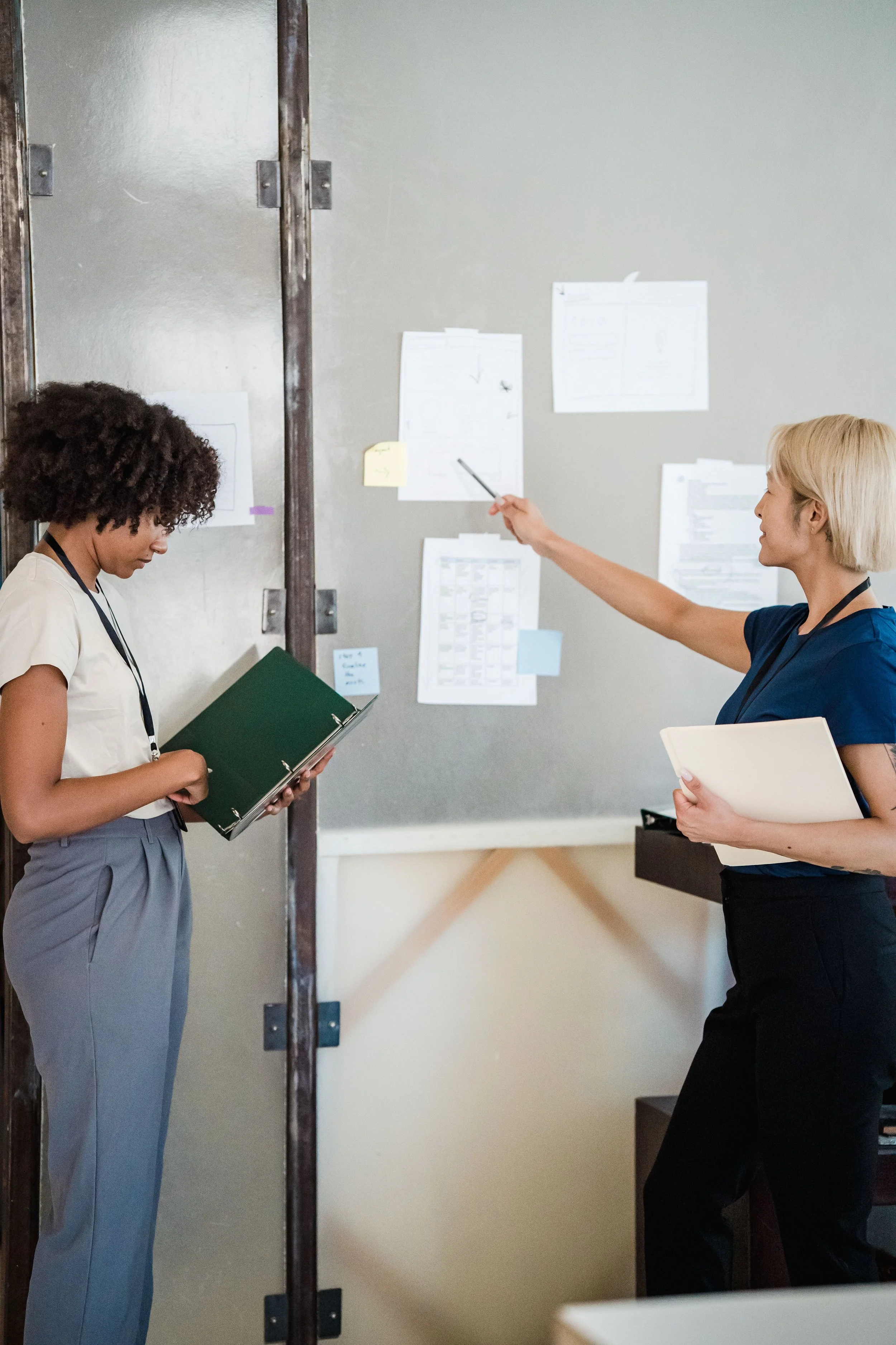 Two women standing in front of a wall with papers attached, one woman with curly hair holding a binder, and the other with blonde hair holding a folder, both engaged in discussion.