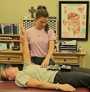 A woman performing a medical or chiropractic adjustment on a man lying on a treatment table in a clinic or office setting.