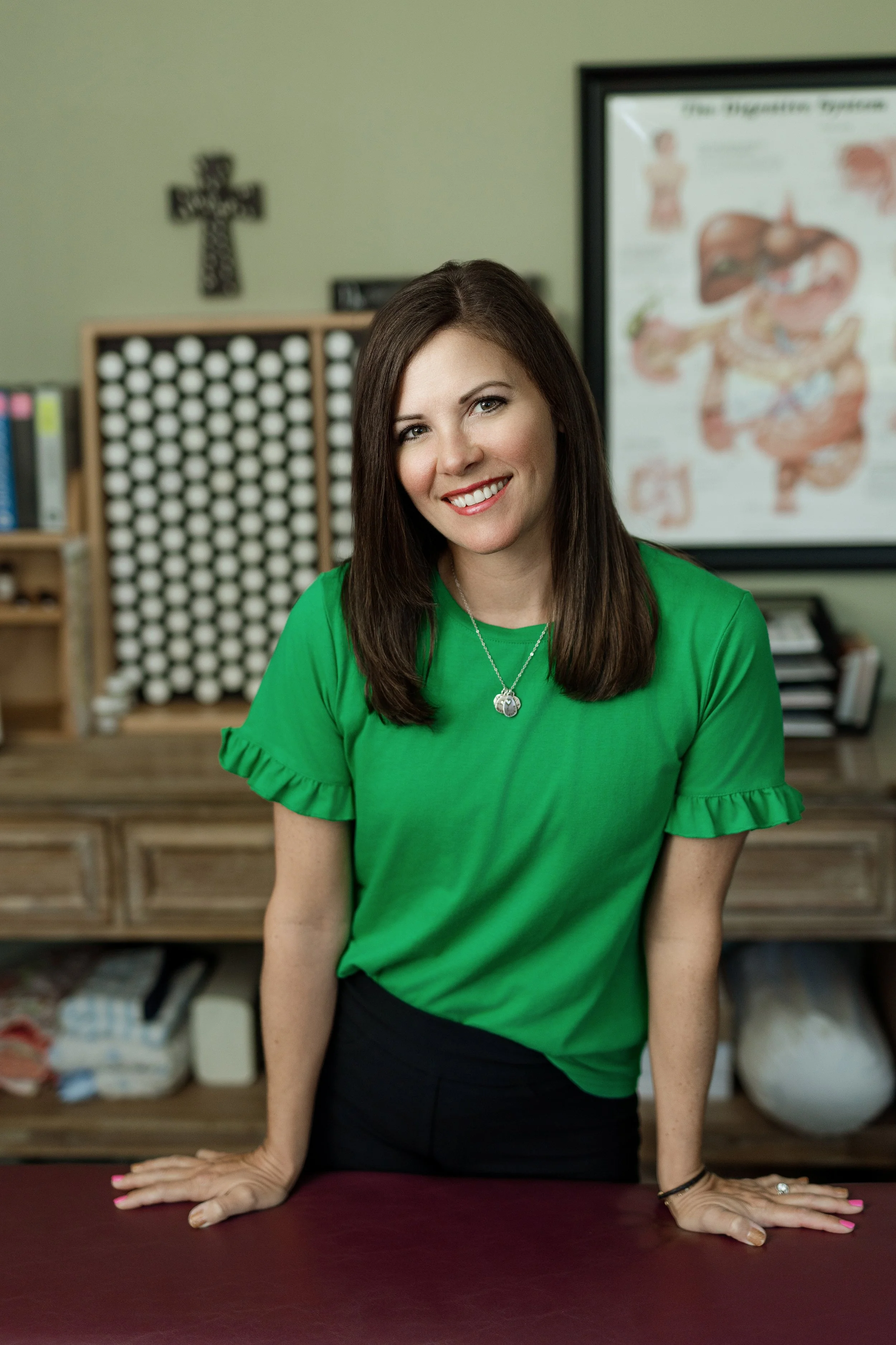 A woman with long brown hair, wearing a green top, standing behind a table with her hands resting on it, smiling in a room with medical posters and a cross on the wall.