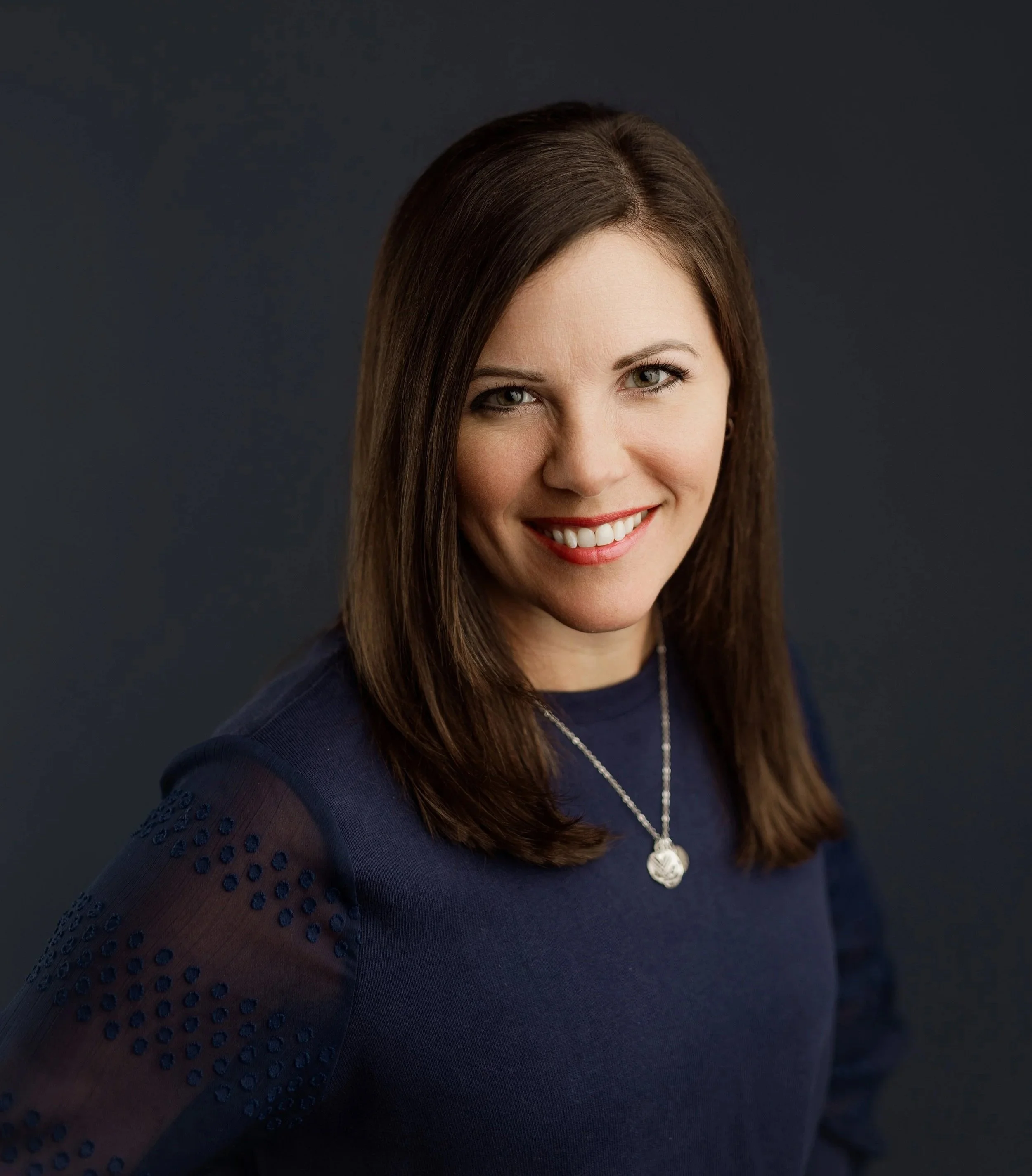 A woman with brown hair, blue eyes, and a bright smile wearing a dark blue top with eyelet detail on the sleeves, a silver necklace with a pendant, and red lipstick.