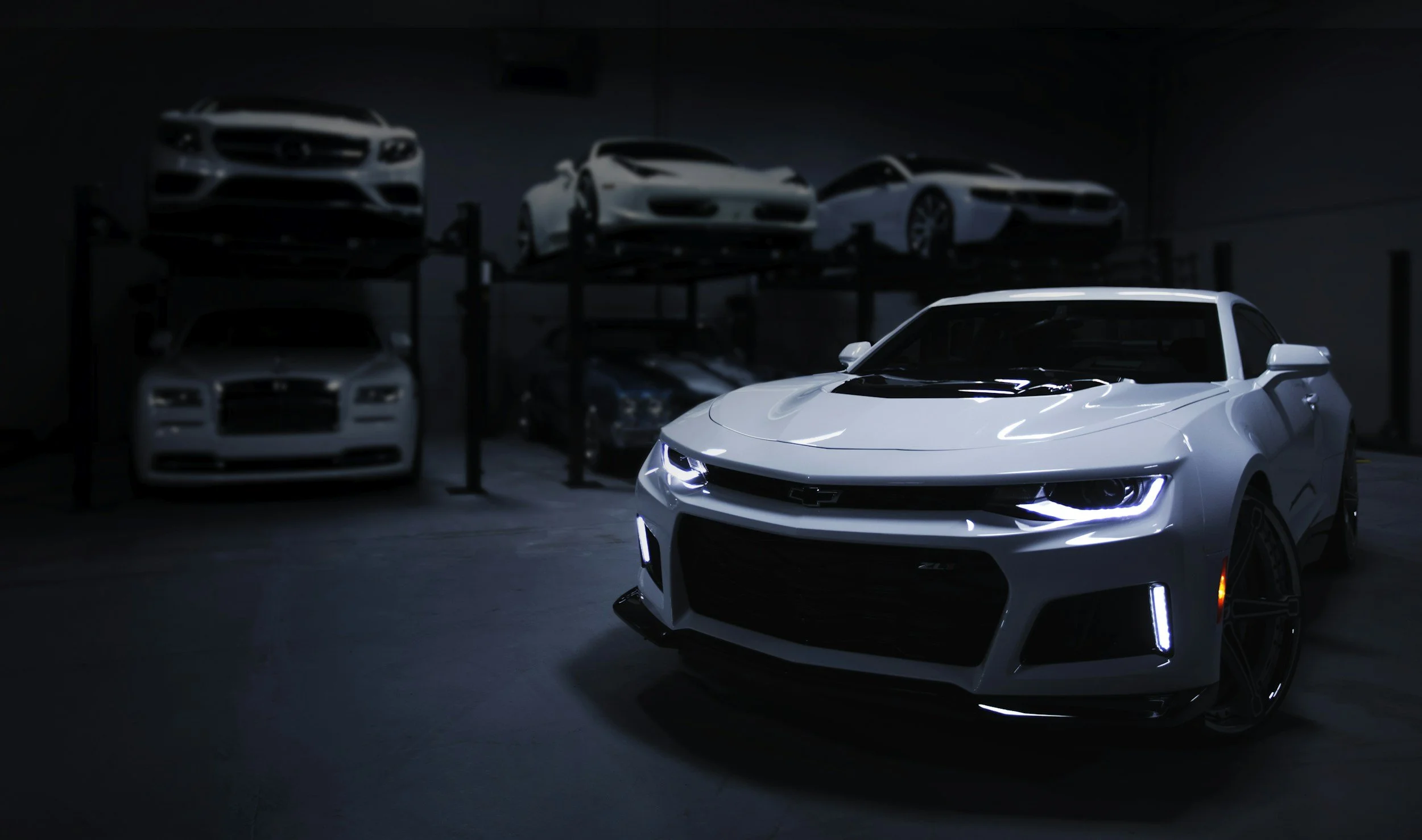 A white Chevrolet sports car with illuminated headlights in a dark garage, with other sleek cars displayed on raised platforms in the background.
