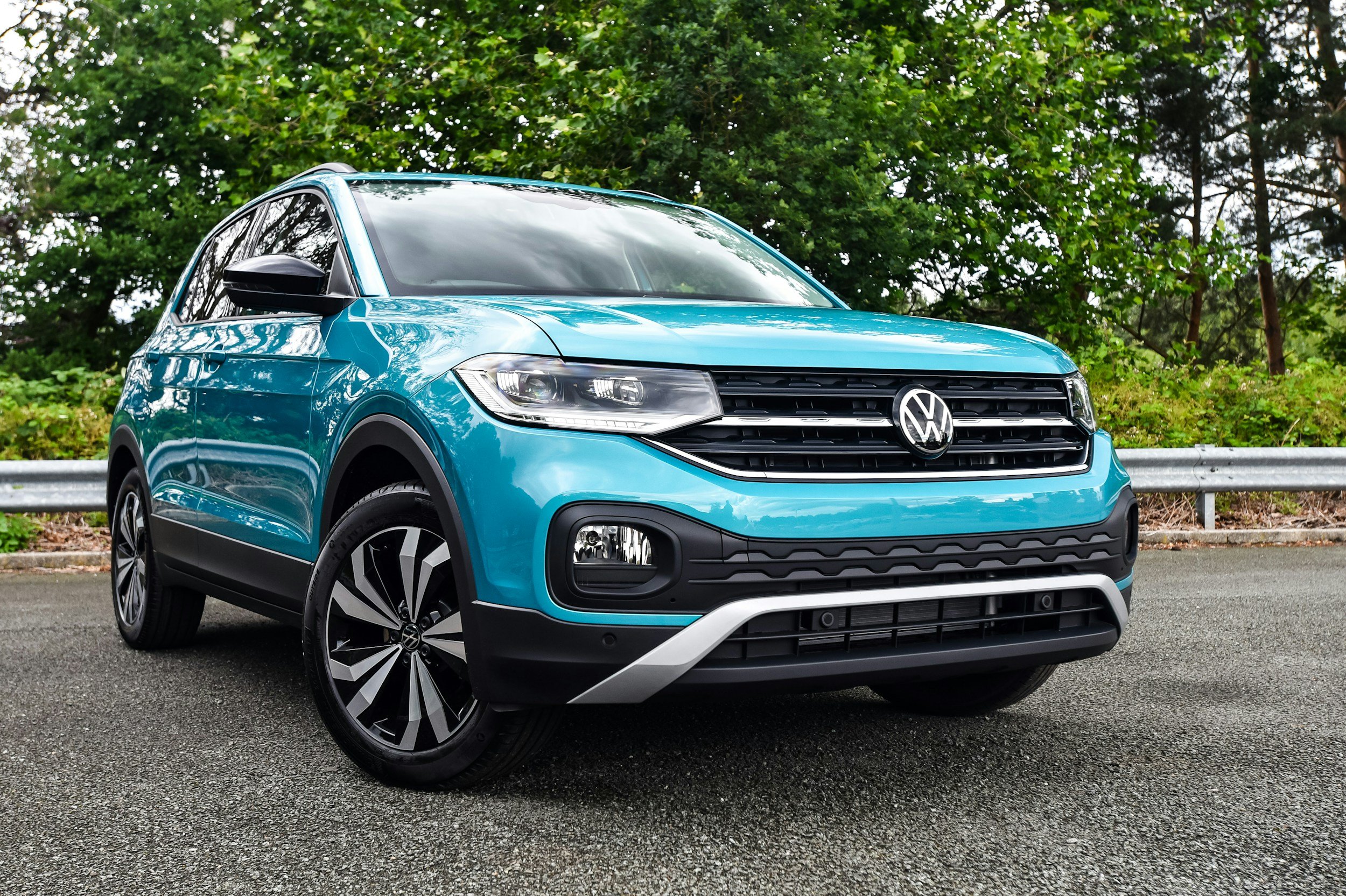 A blue Volkswagen compact SUV parked on a paved surface with greenery and trees in the background.