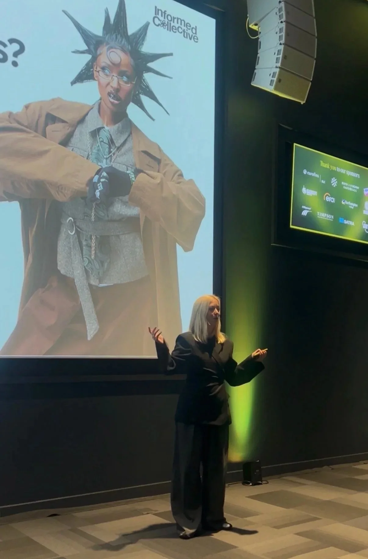 A woman with blonde hair and glasses giving a presentation in front of a large screen displaying a graphic of a person with spiky hair and glasses, with the words "Informed Collective." The presenter is gesturing with her hands, and the scene is illuminated with green lighting.