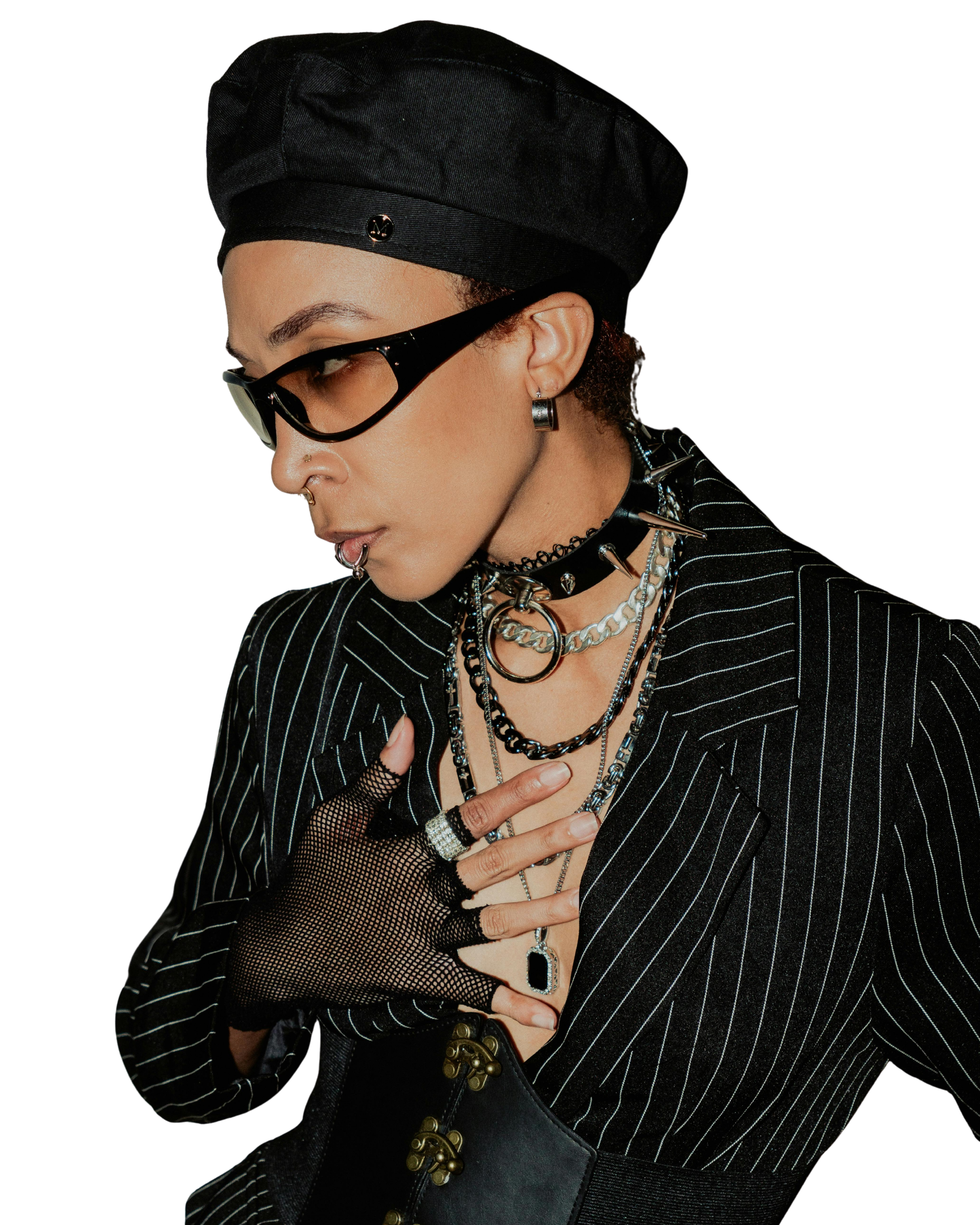 A person with short curly hair and multiple piercings, wearing black sunglasses, a black beret, a striped blazer, and layered jewelry, including chains and rings.