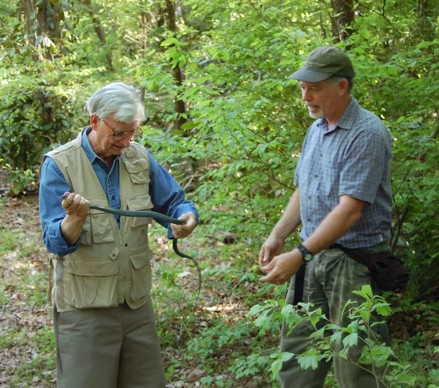 Two men in a forest holding a snake, one demonstrating and the other observing.
