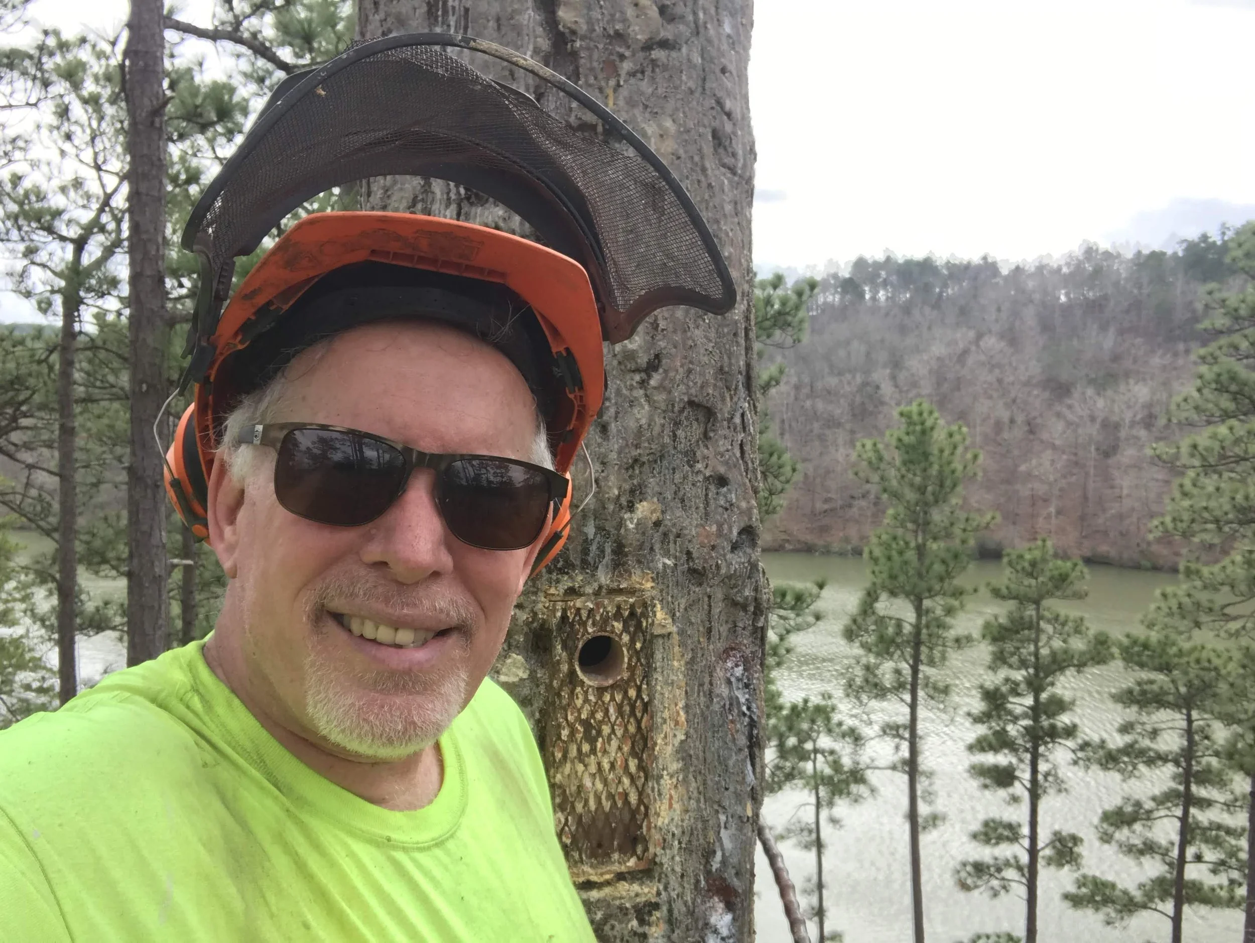 Close-up of a smiling man with sunglasses wearing a bright yellow shirt, standing outdoors near a tree with a landscape of trees and water in the background.
