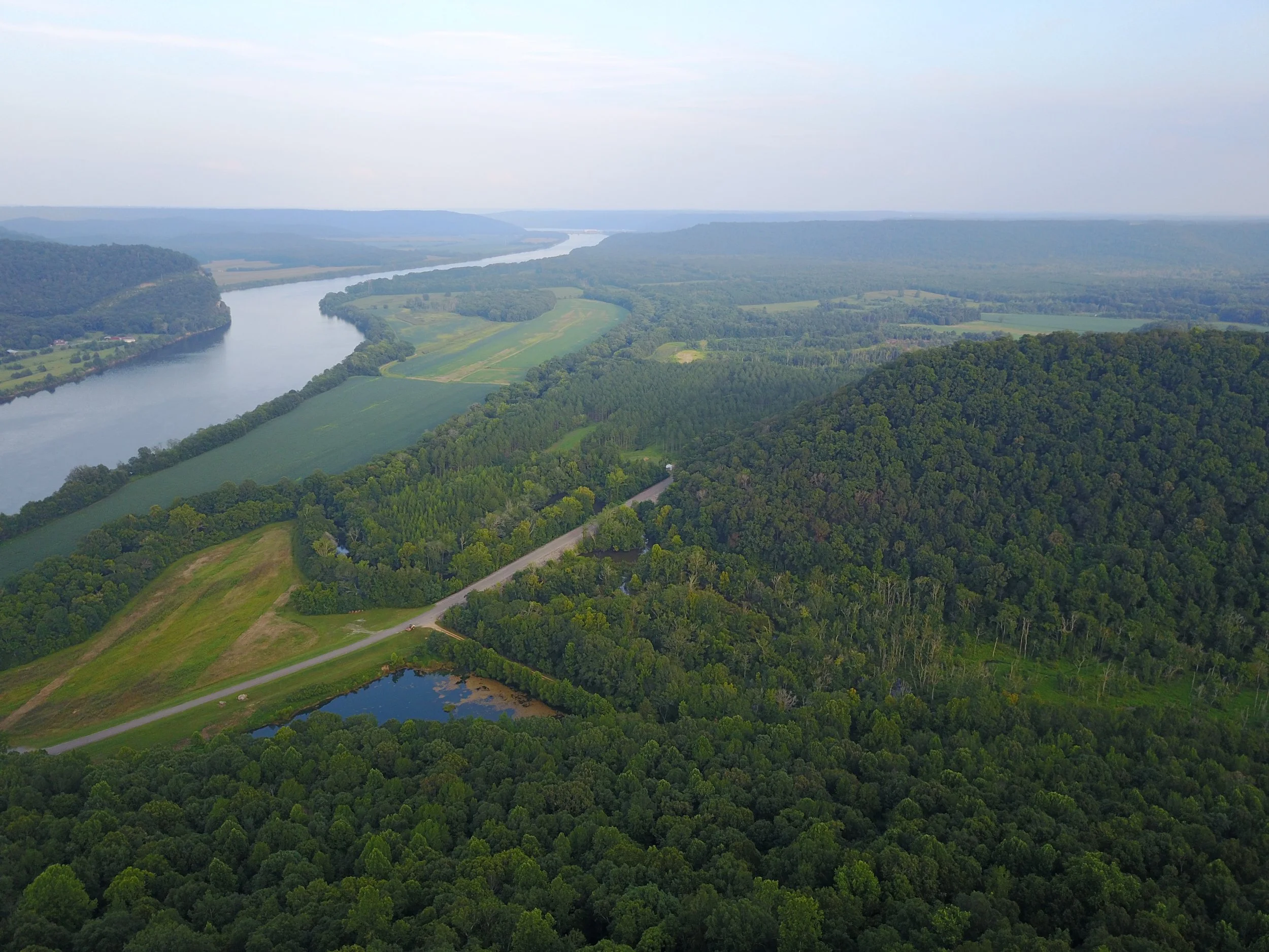 Aerial view of a winding river flowing through lush green forested hills and farmland, with more hills visible in the distance under a partly cloudy sky.
