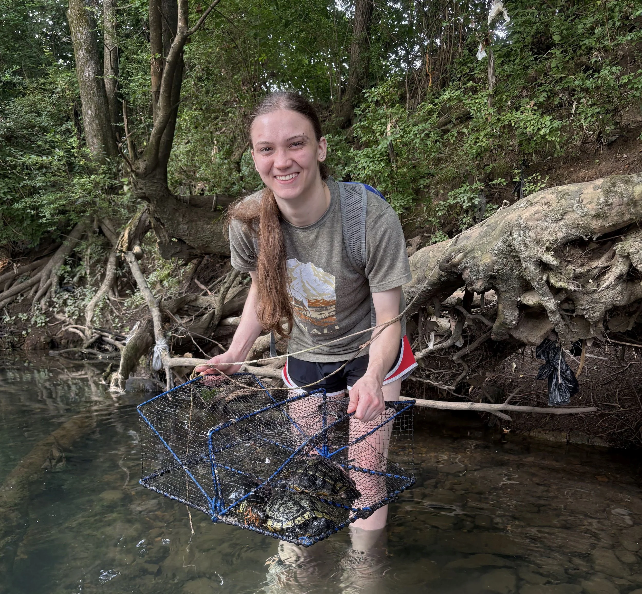 A young woman with long hair, wearing a t-shirt and shorts, stands in a shallow river holding a fishing net. She is smiling, surrounded by dense green trees and bushes along the riverbank.