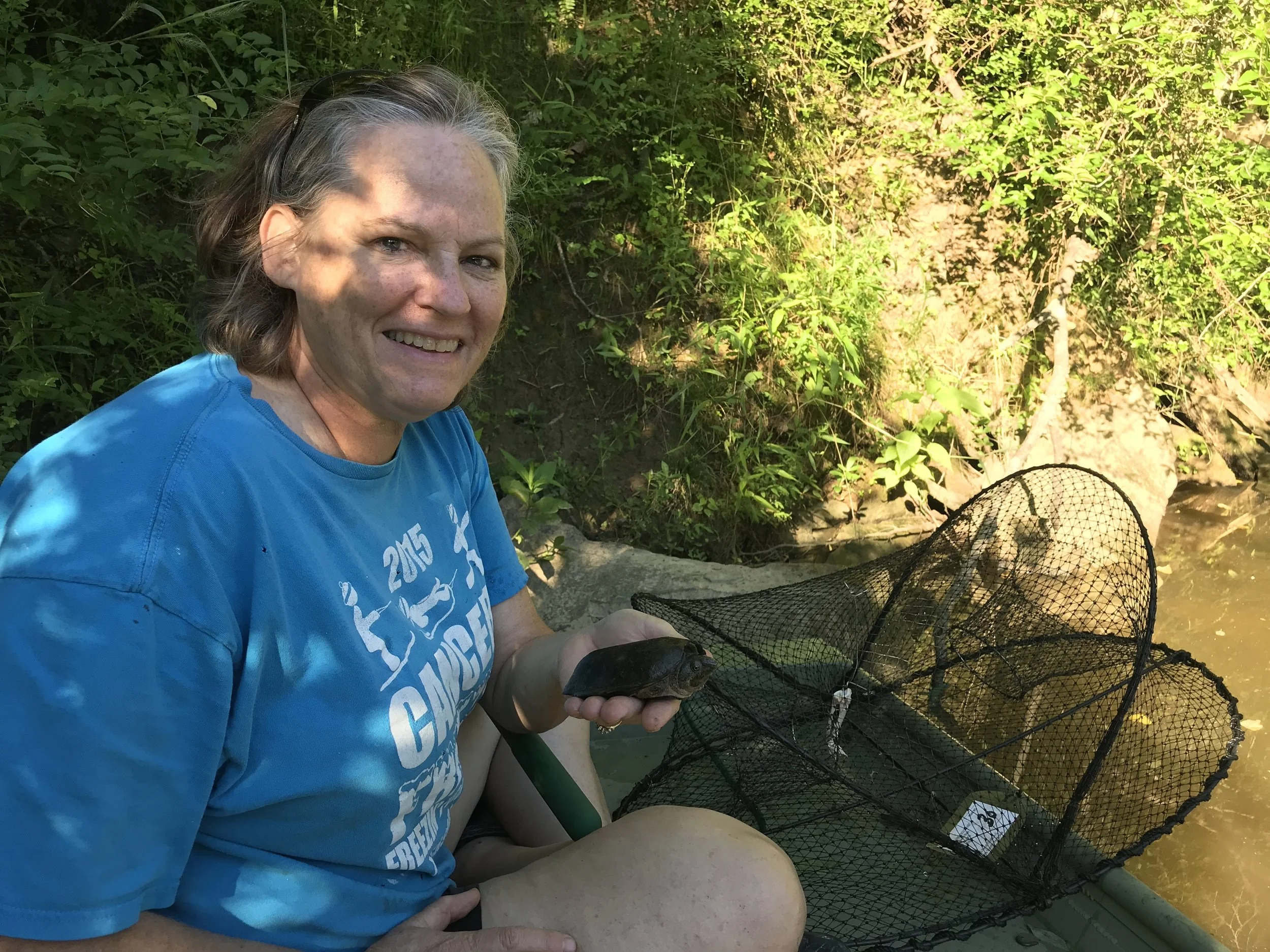 A woman with gray hair smiling while holding a fish in her hand by a river, surrounded by green foliage, during daytime.