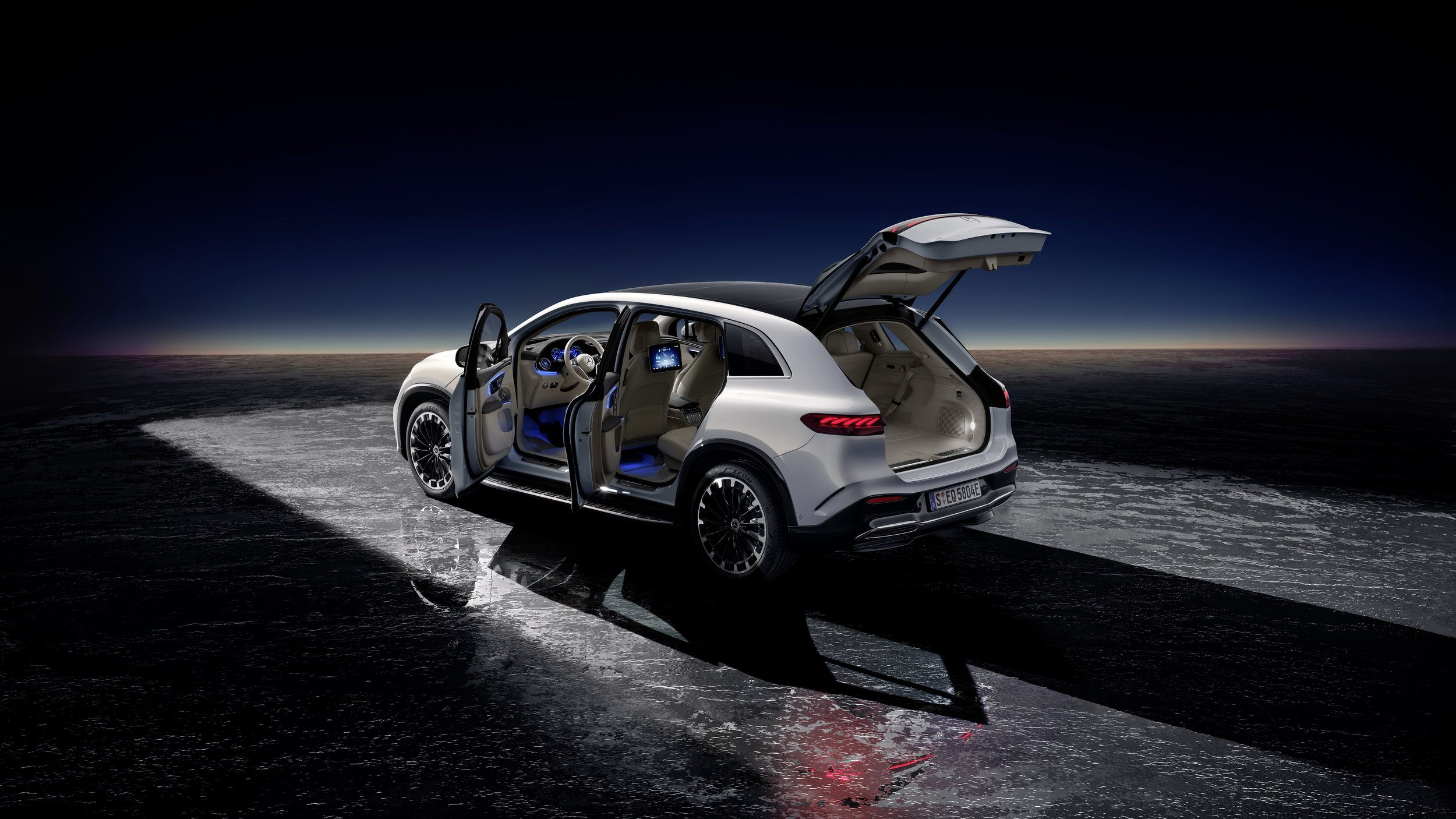 A white Mercedes-Benz EQS SUV with open front and rear doors, showing illuminated interior and dashboard, parked on a reflective surface under a dark sky at sunset or night.