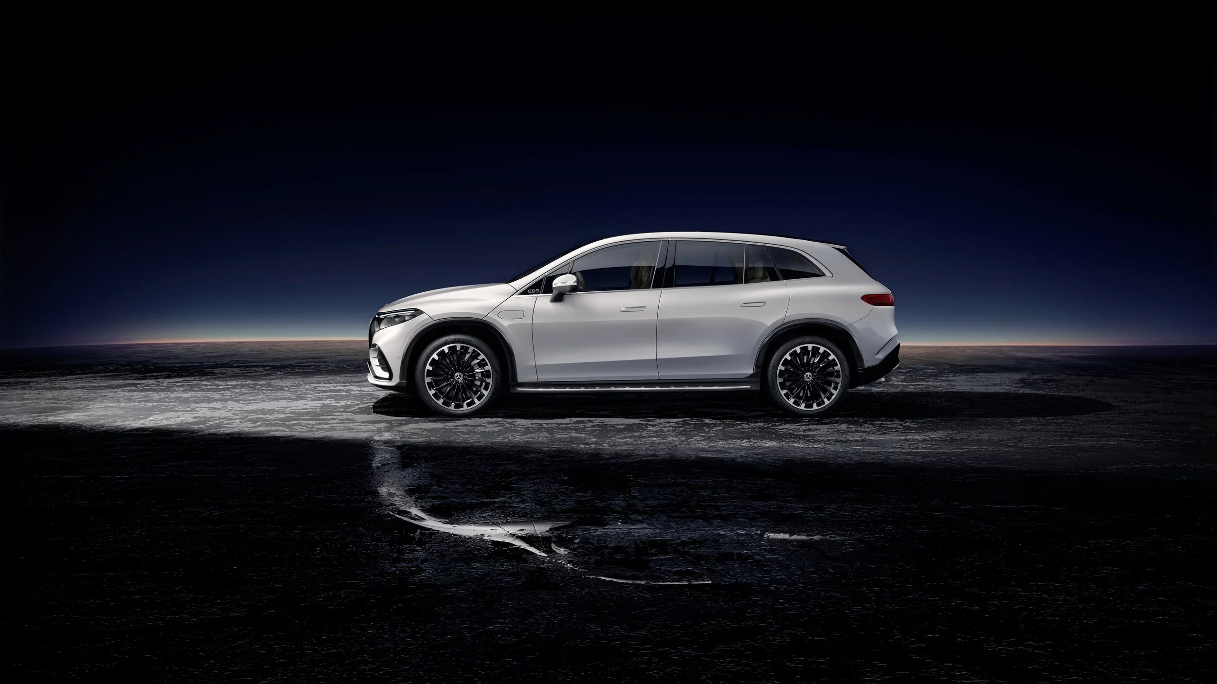 A white Mercedes-Benz EQS SUV parked on a reflective wet surface under a dark sky with a sunset horizon