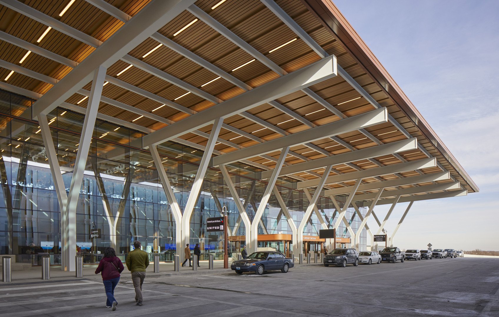 Exterior of a modern airport terminal with large glass windows and a distinctive angled roof structure, with cars parked in front and people walking nearby.