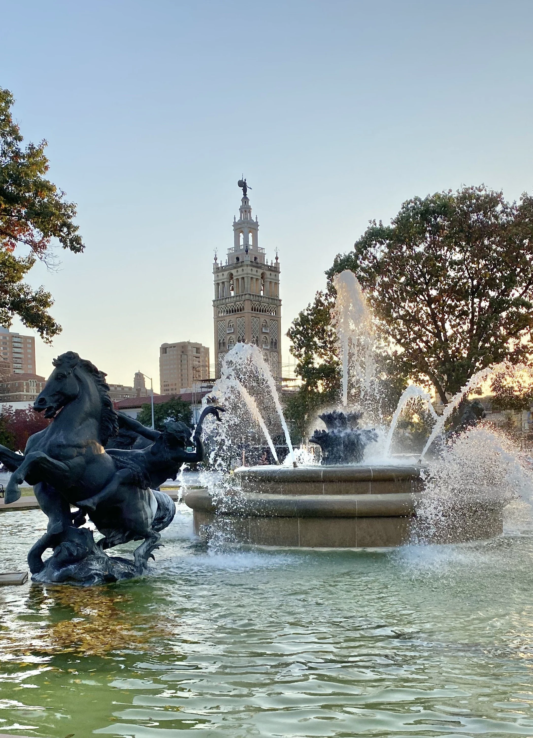 Fountain with bronze horse sculptures in a city park, water spraying from the fountain, with a clock tower and trees in the background, during late afternoon or early evening.
