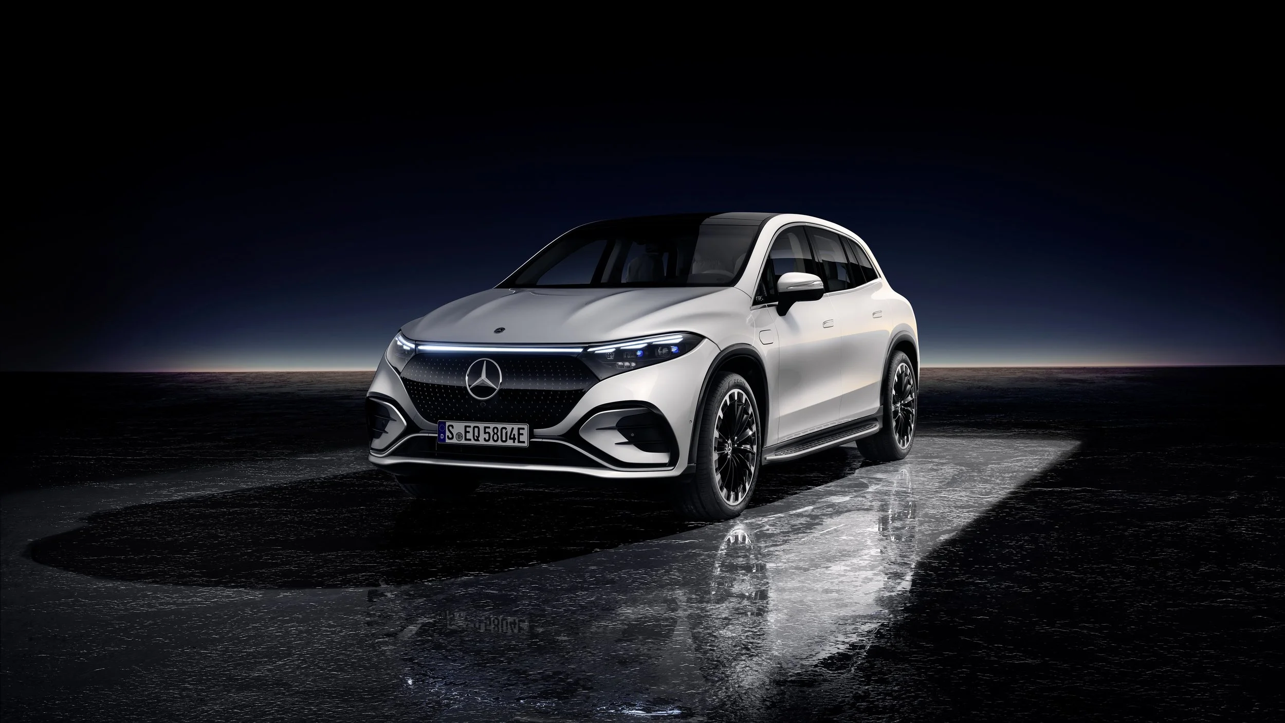 A white Mercedes-Benz EQS SUV parked on a reflective surface at night with a dark sky and horizon in the background.