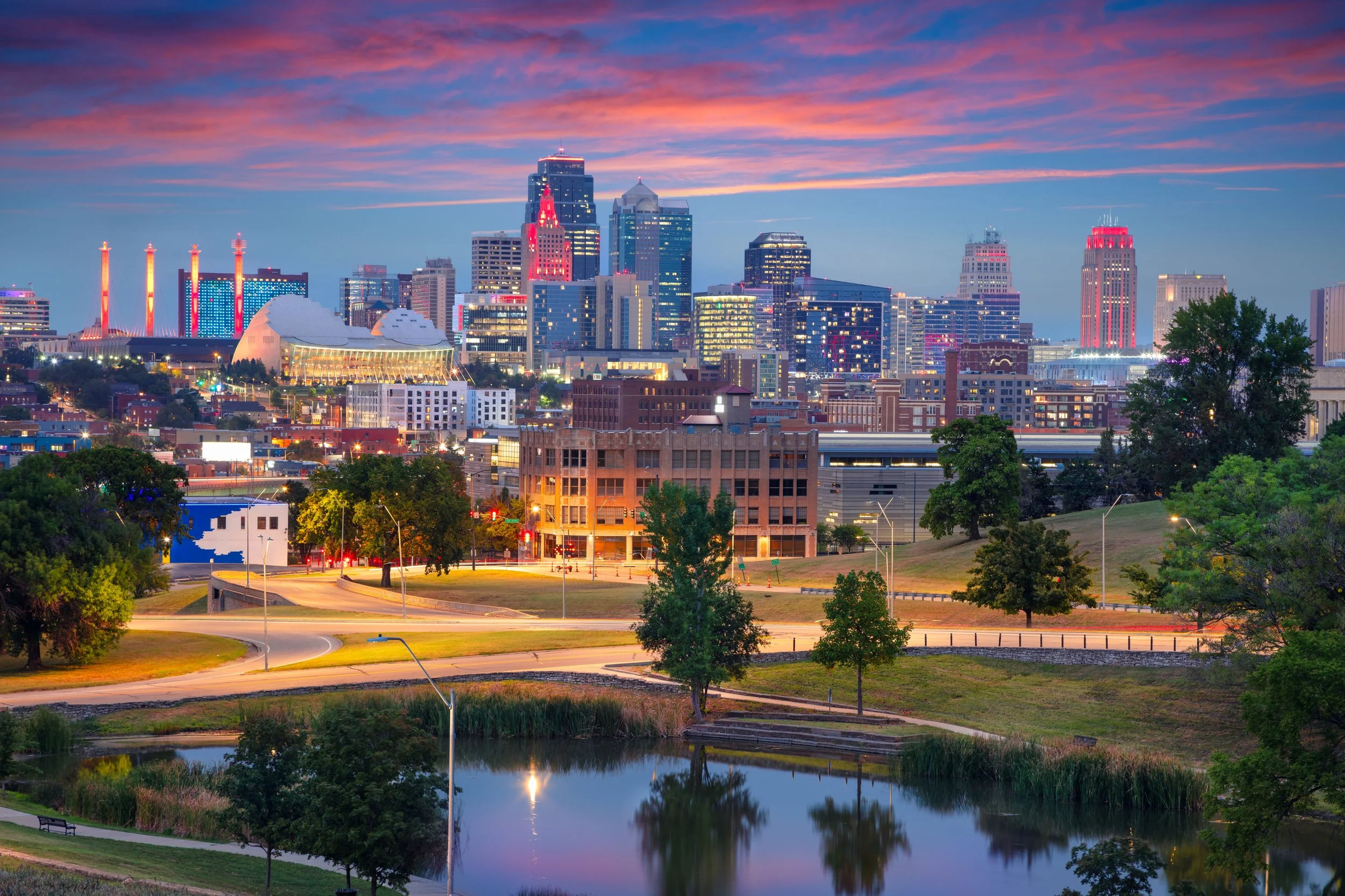 Nighttime city skyline with illuminated skyscrapers, a body of water in the foreground reflecting the city lights, and a colorful sky with pink and purple clouds.