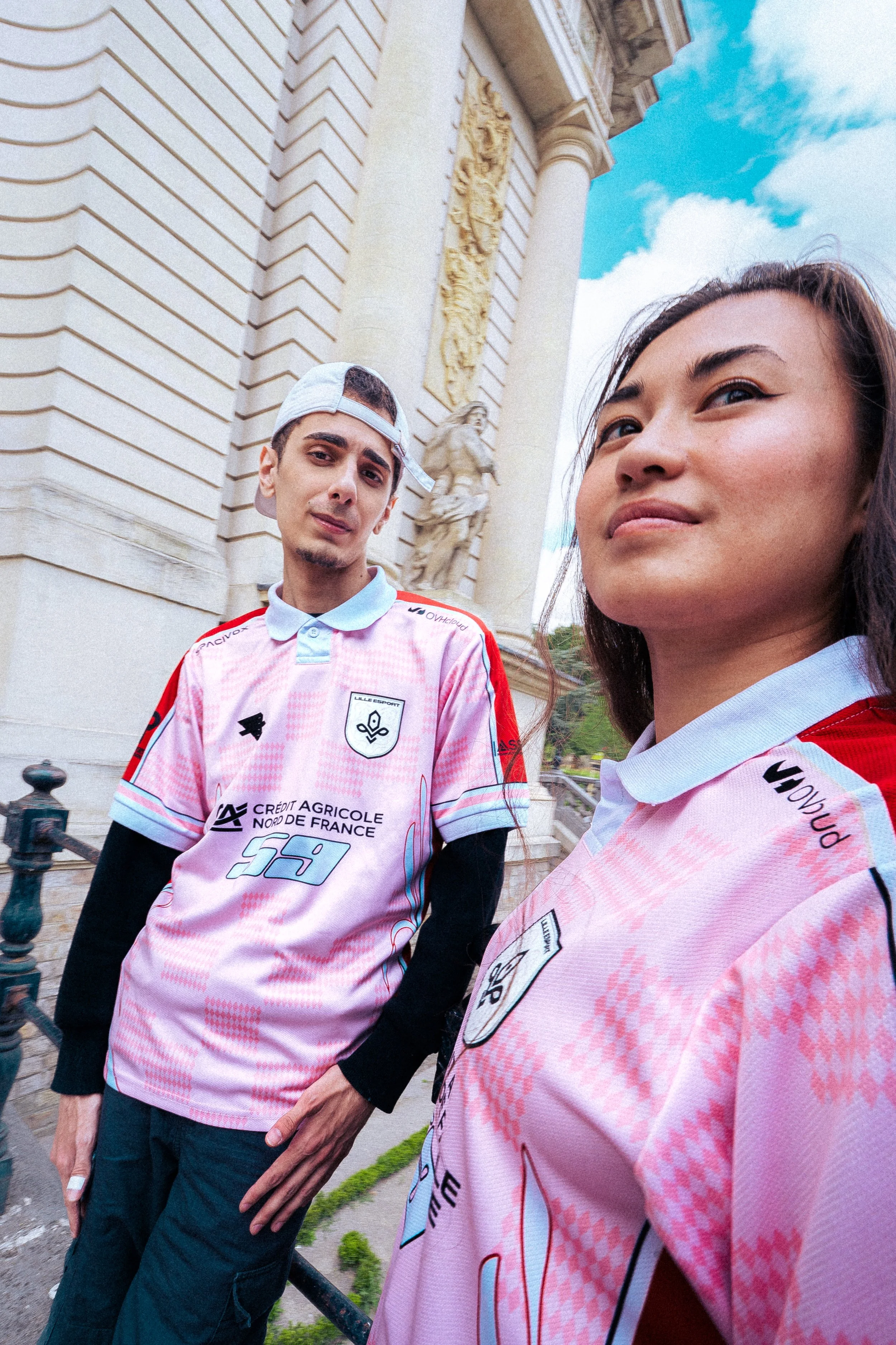 Deux jeunes adultes portent des maillots de football rose avec des logos et des sponsors et prennent un selfie devant un bâtiment historique en pierre blanche. Le ciel est partiellement nuageux.