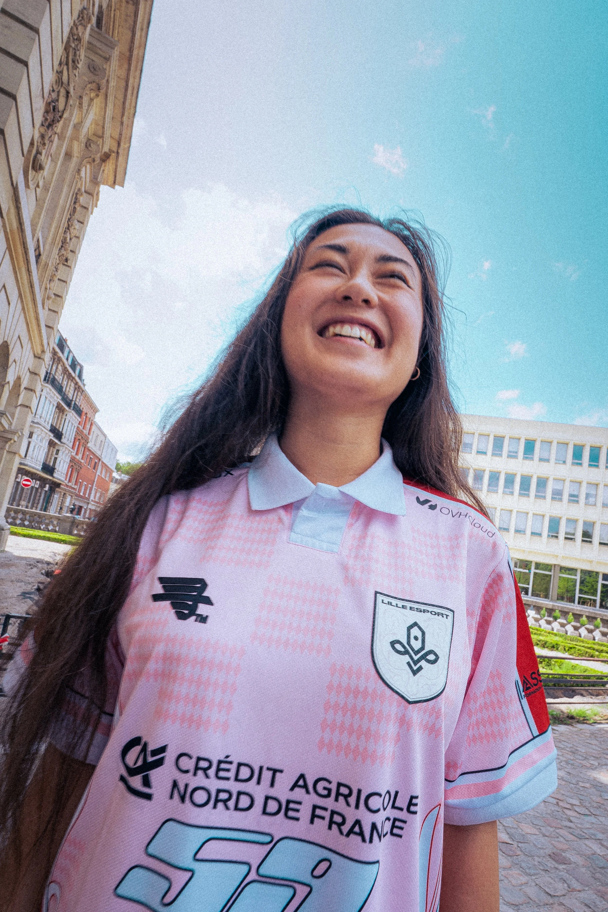 Jeune femme souriante portant un maillot de sport rose et blanc avec le logo de Lille OSC, debout à l'extérieur sous un ciel bleu, avec des bâtiments classiques en arrière-plan.