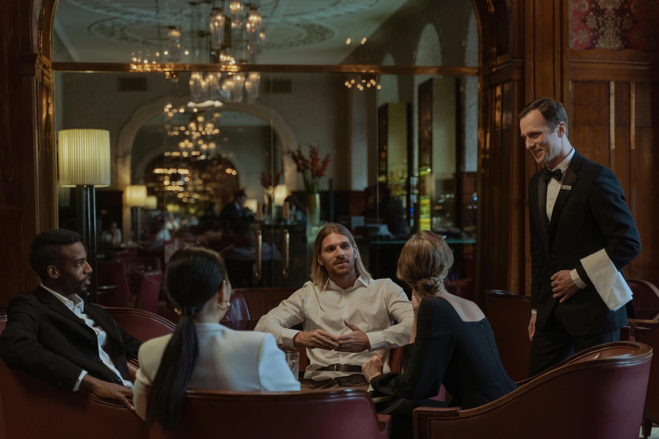 A waiter in a tuxedo serving a group of six people in a restaurant with elegant wooden decor, large mirror, chandeliers, and warm lighting.