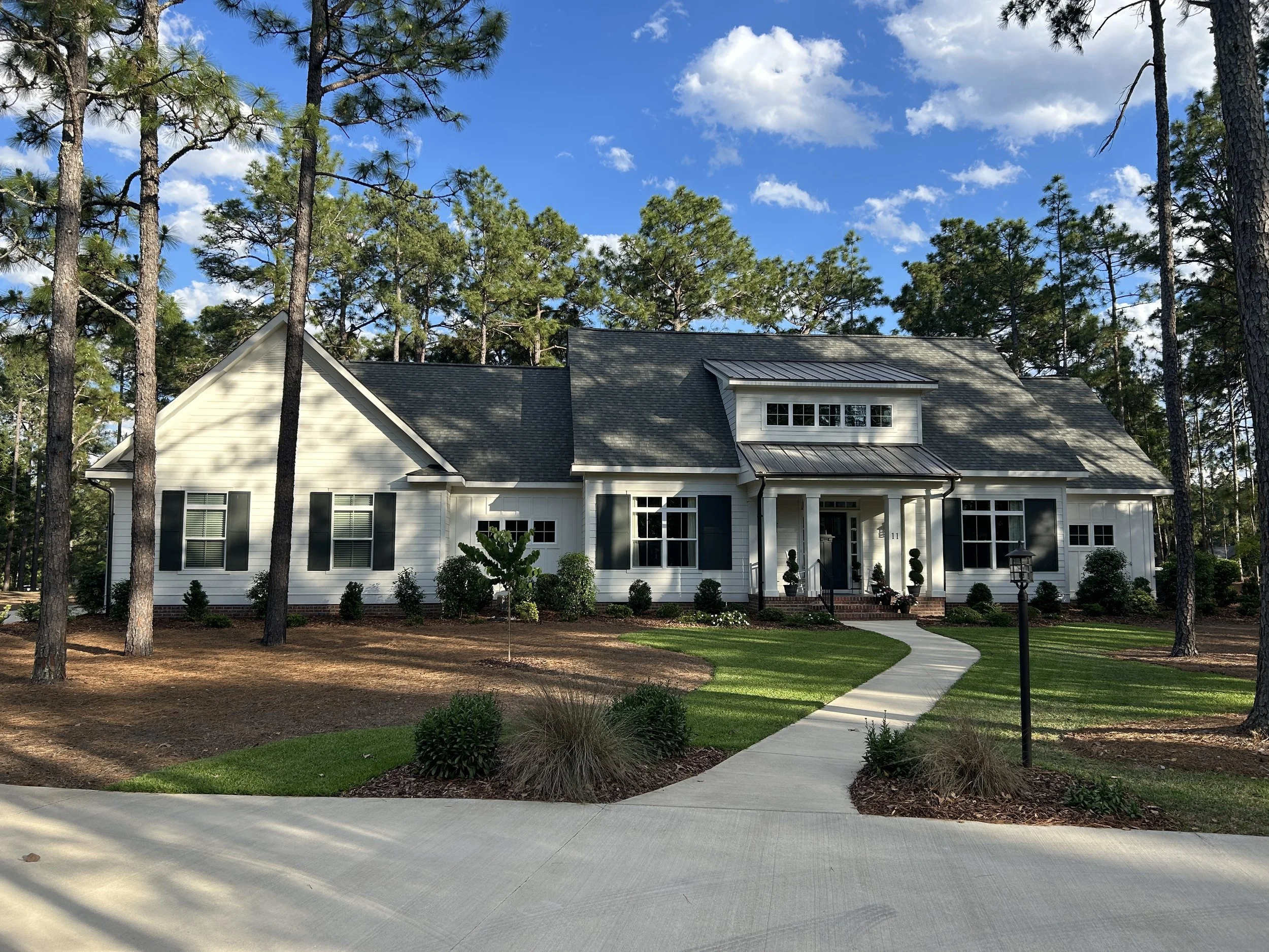 A classic white house with dark green shutters and am metal roof, surrounded by tall pine trees and a well-maintained garden under a blue sky with some clouds.