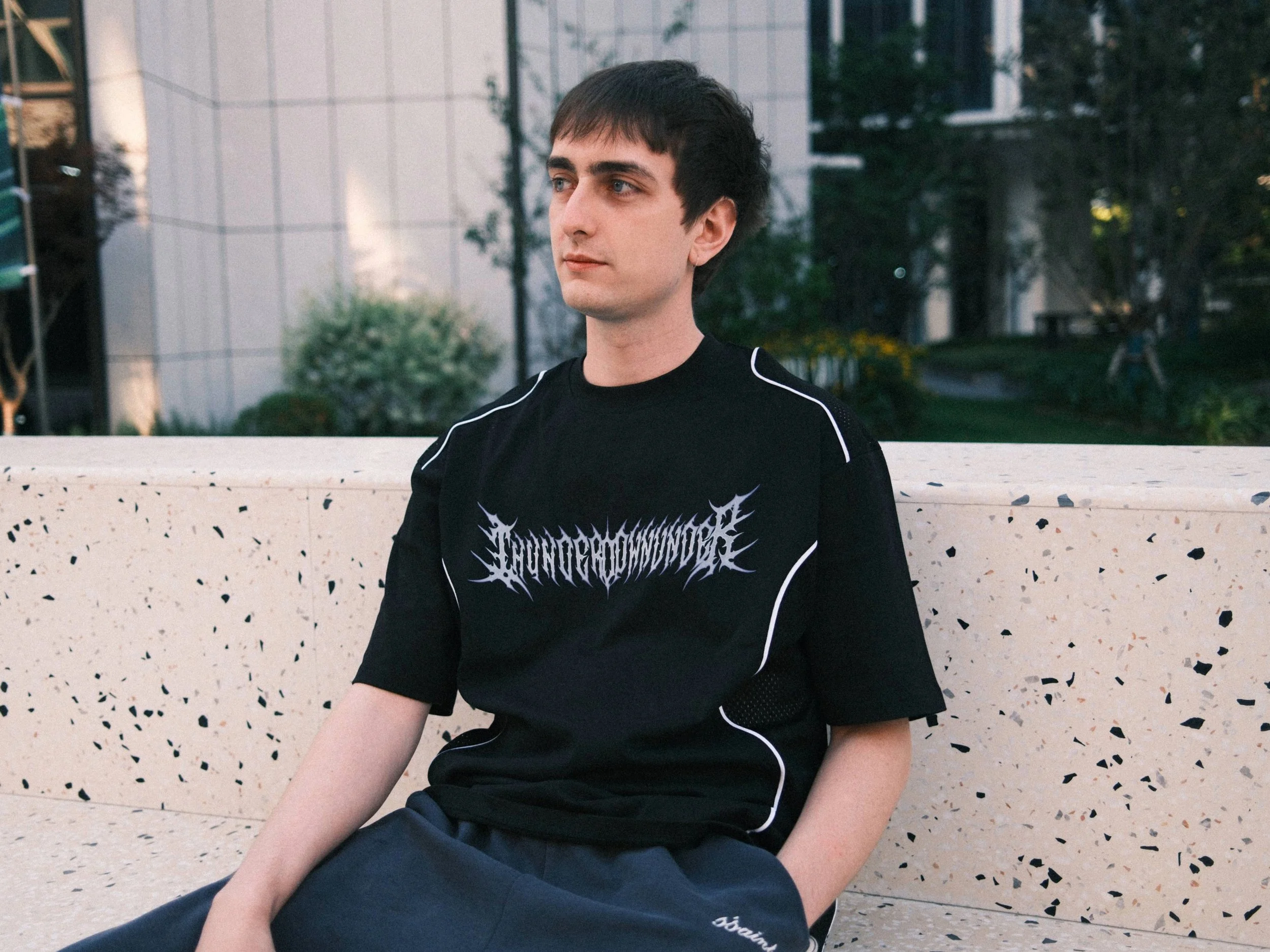 A young man with dark hair and blue eyes sitting on a bench outdoors, dressed in a black athletic shirt with white graphics and dark shorts.