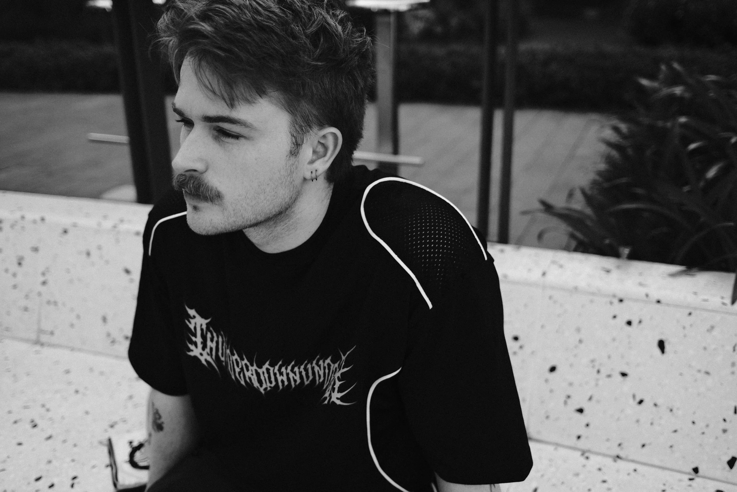 A young man with dark, wavy hair, a mustache, and earrings sitting outdoors near a railing and plants, in black and white.