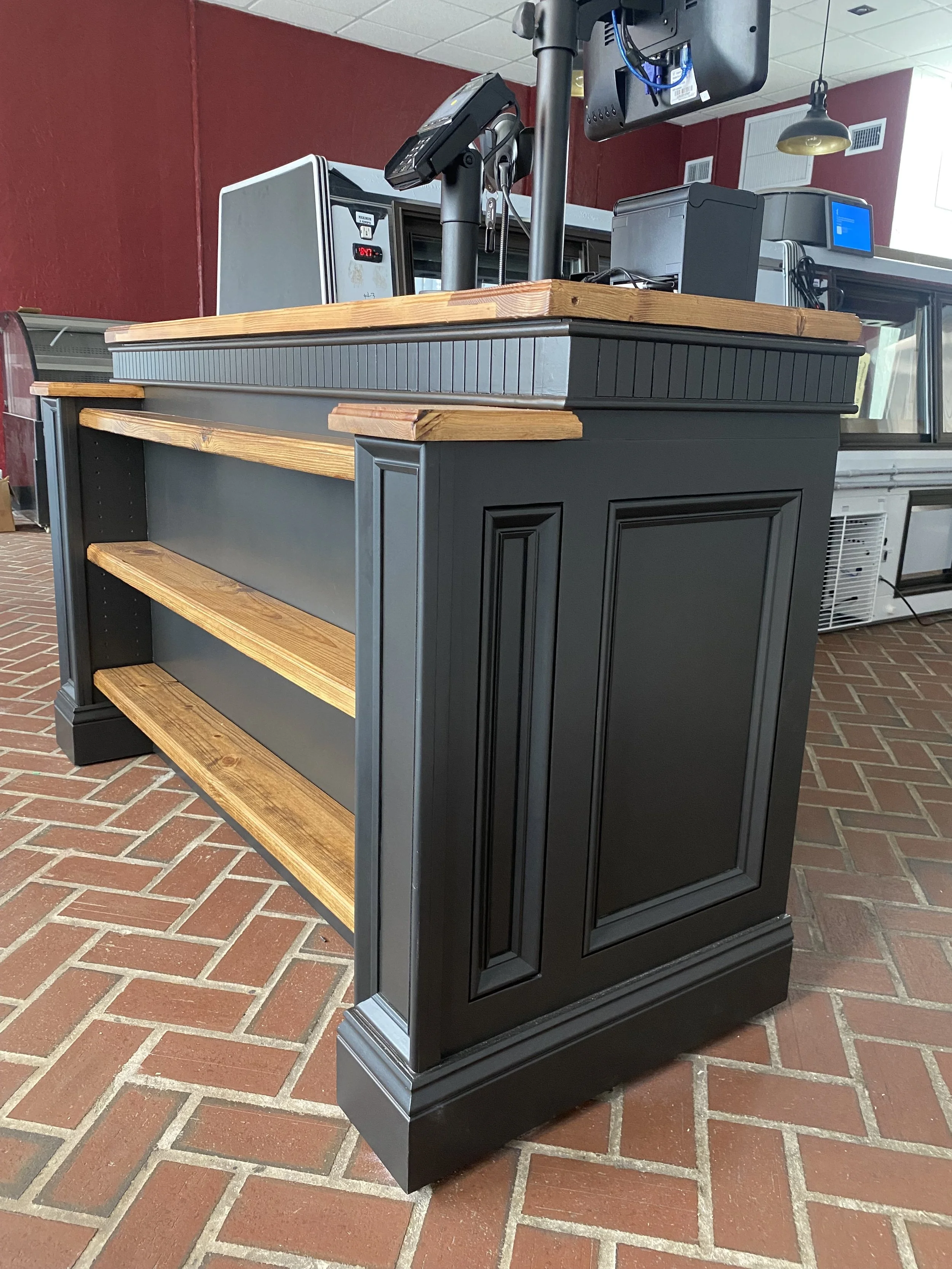 A black and wooden retail or restaurant counter with shelving, situated on a brick floor, with a red wall in the background.