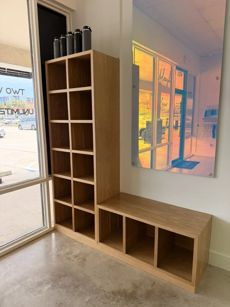 Empty wooden cubby shelf unit near a window, with a few black water bottles on top, and a colorful abstract wall art piece next to it.