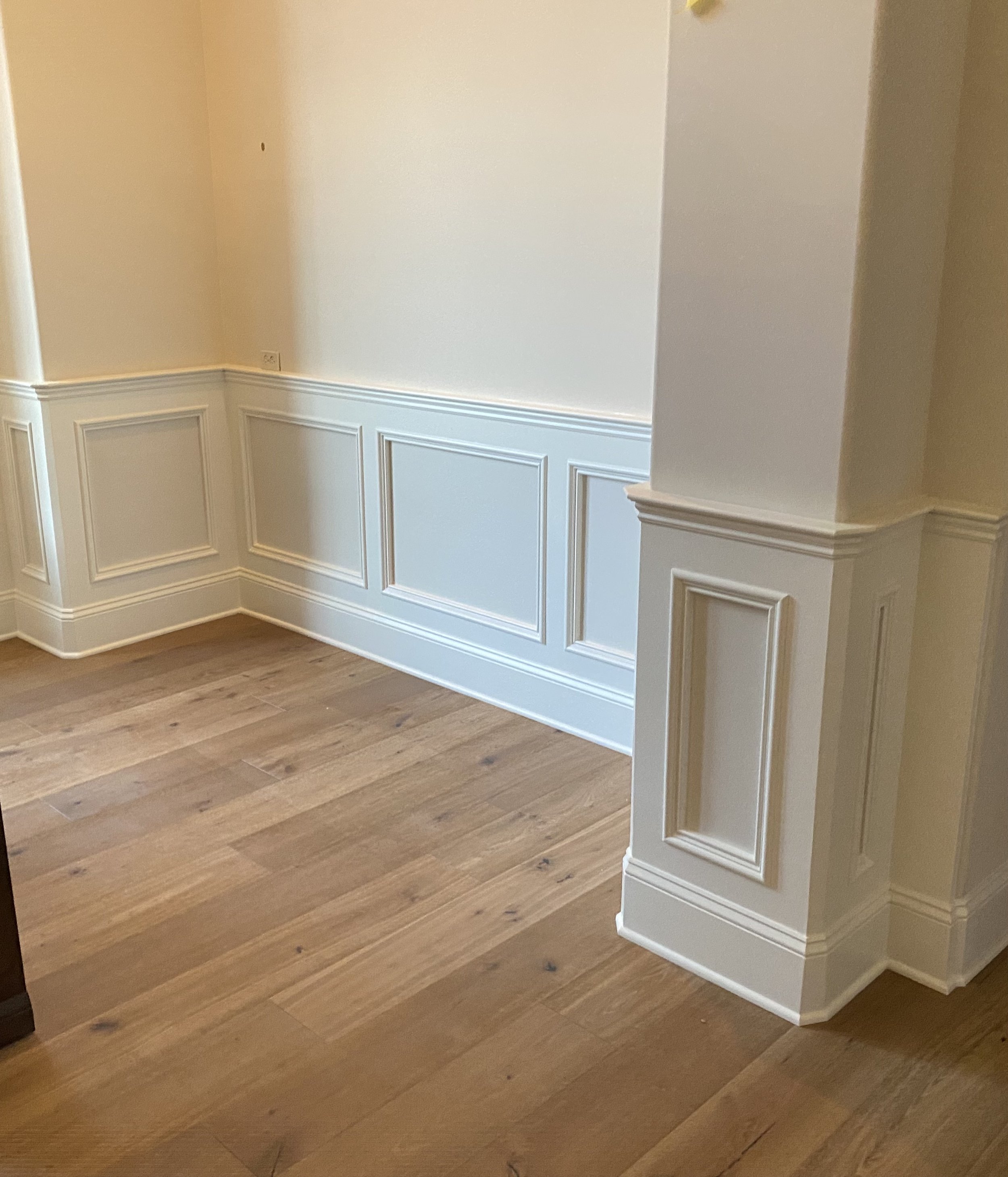Empty corner of a room with beige walls, white wainscoting, and hardwood floors.