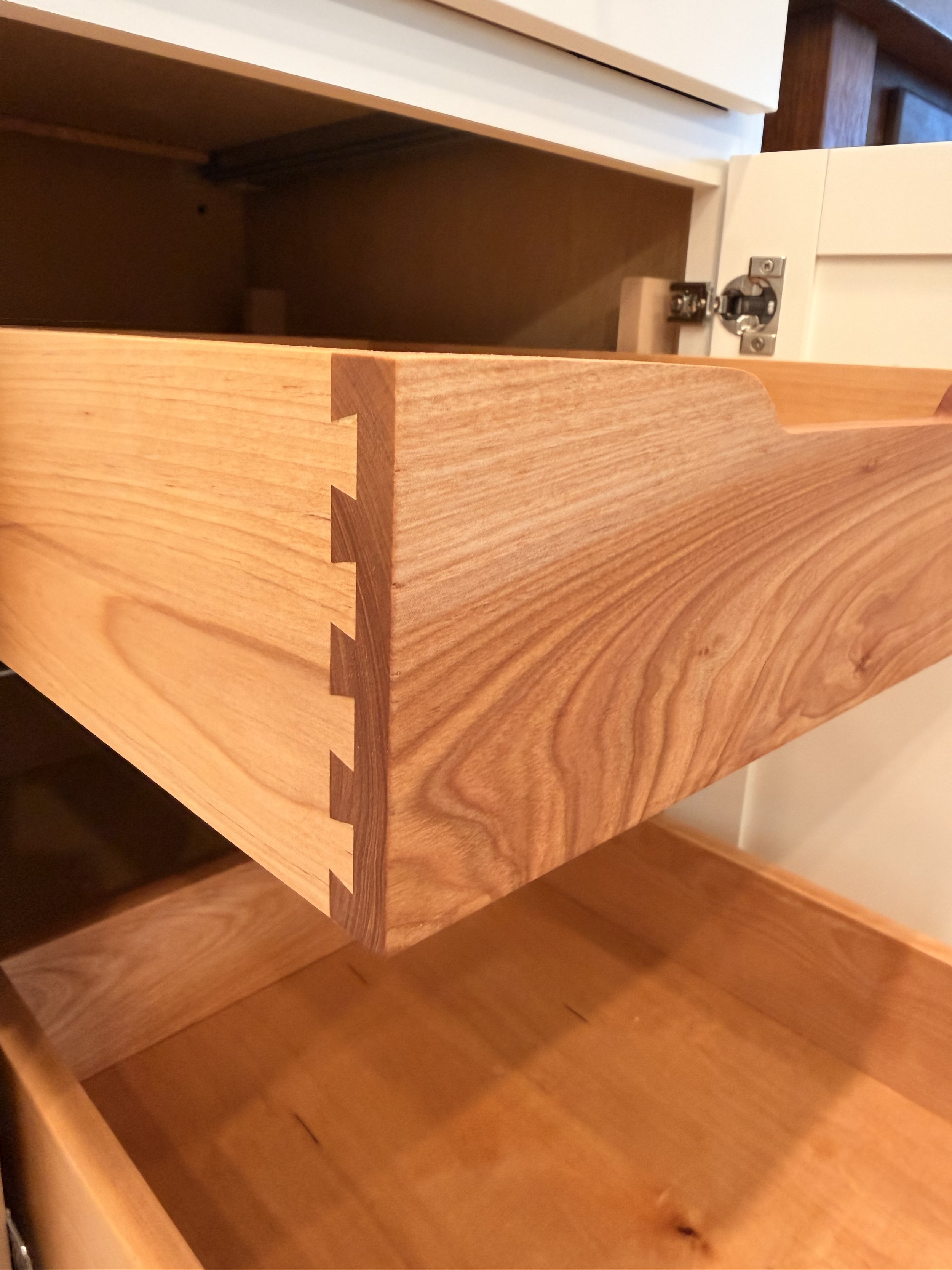 A wooden drawer with dovetail joints on the front, partially open, showing dark interior, mounted inside a wooden cabinet.