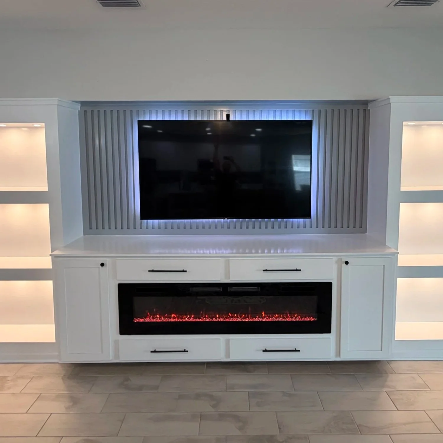 White built-in entertainment center with a wall-mounted flat-screen TV and an electric fireplace below, flanked by illuminated display niches, in a modern living room.